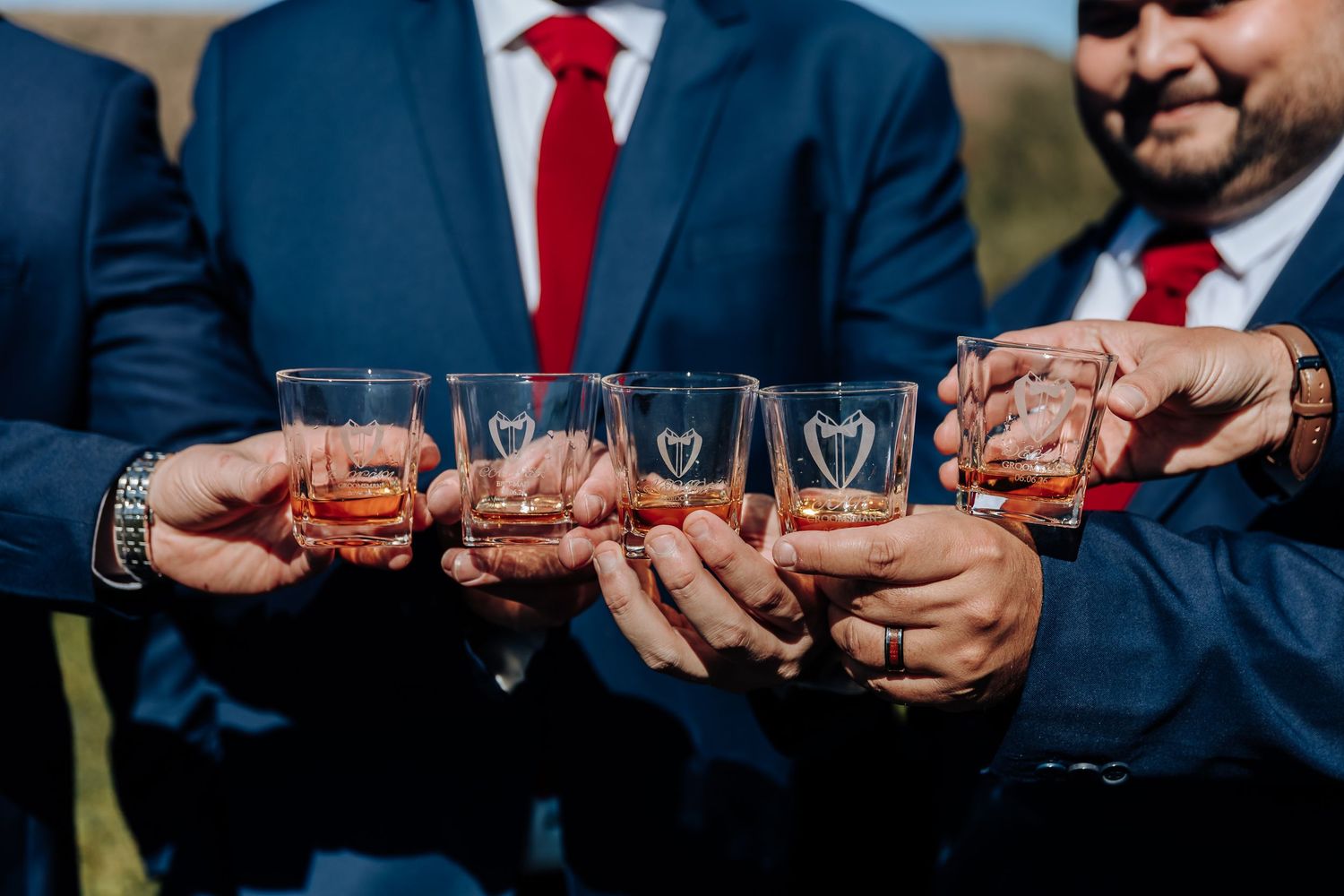 Groomsmen in blue suits and red ties toasting with engraved whiskey glasses at a wedding celebration outdoors.