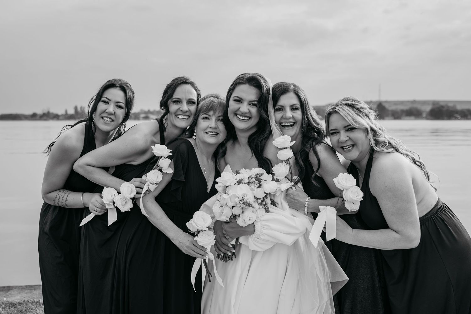 A wedding party in black and white poses together by a lake, with bridesmaids in dark dresses and white bouquets.