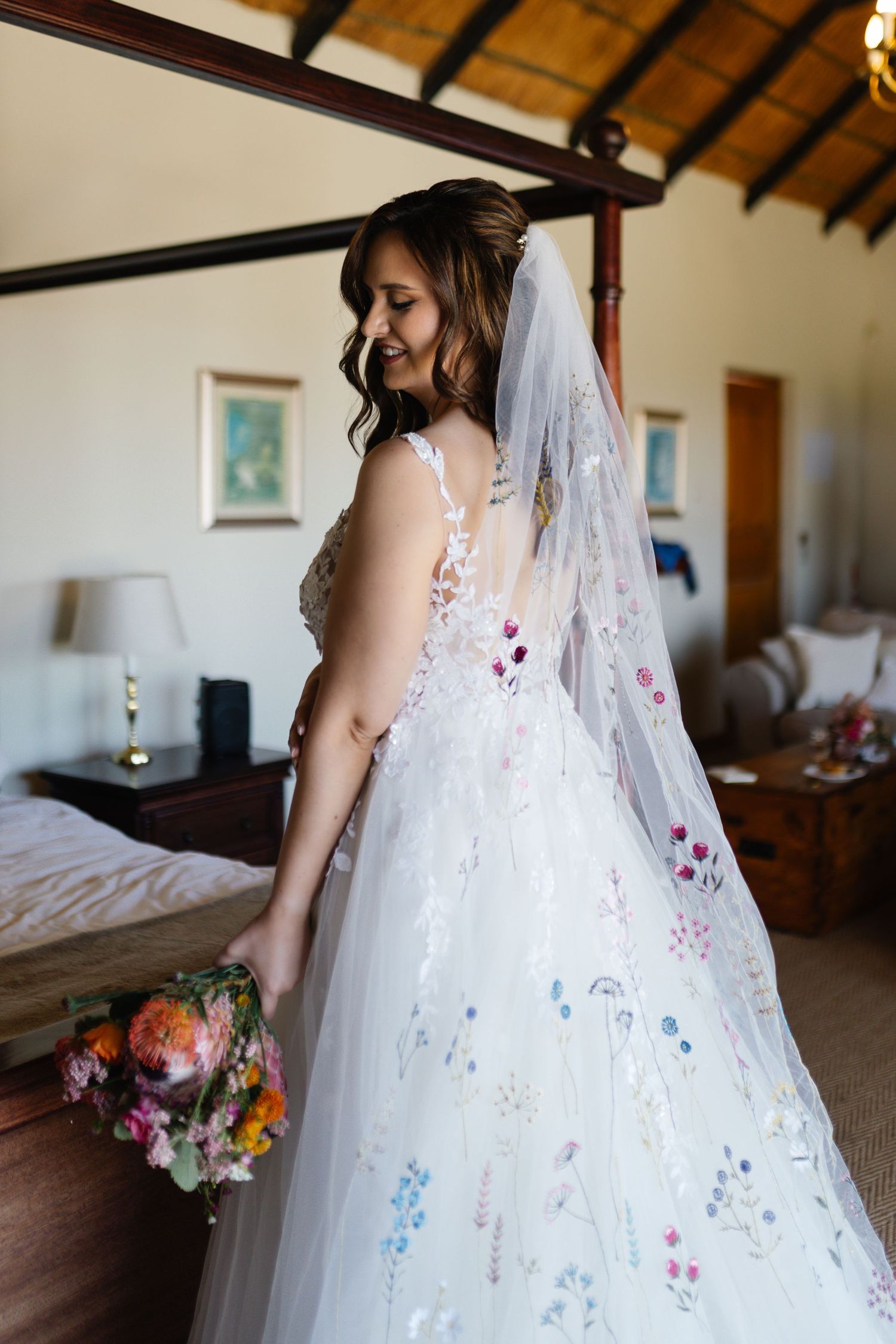 A bride in a delicate white gown with floral embellishments and veil holds a colorful bouquet in a rustic room.