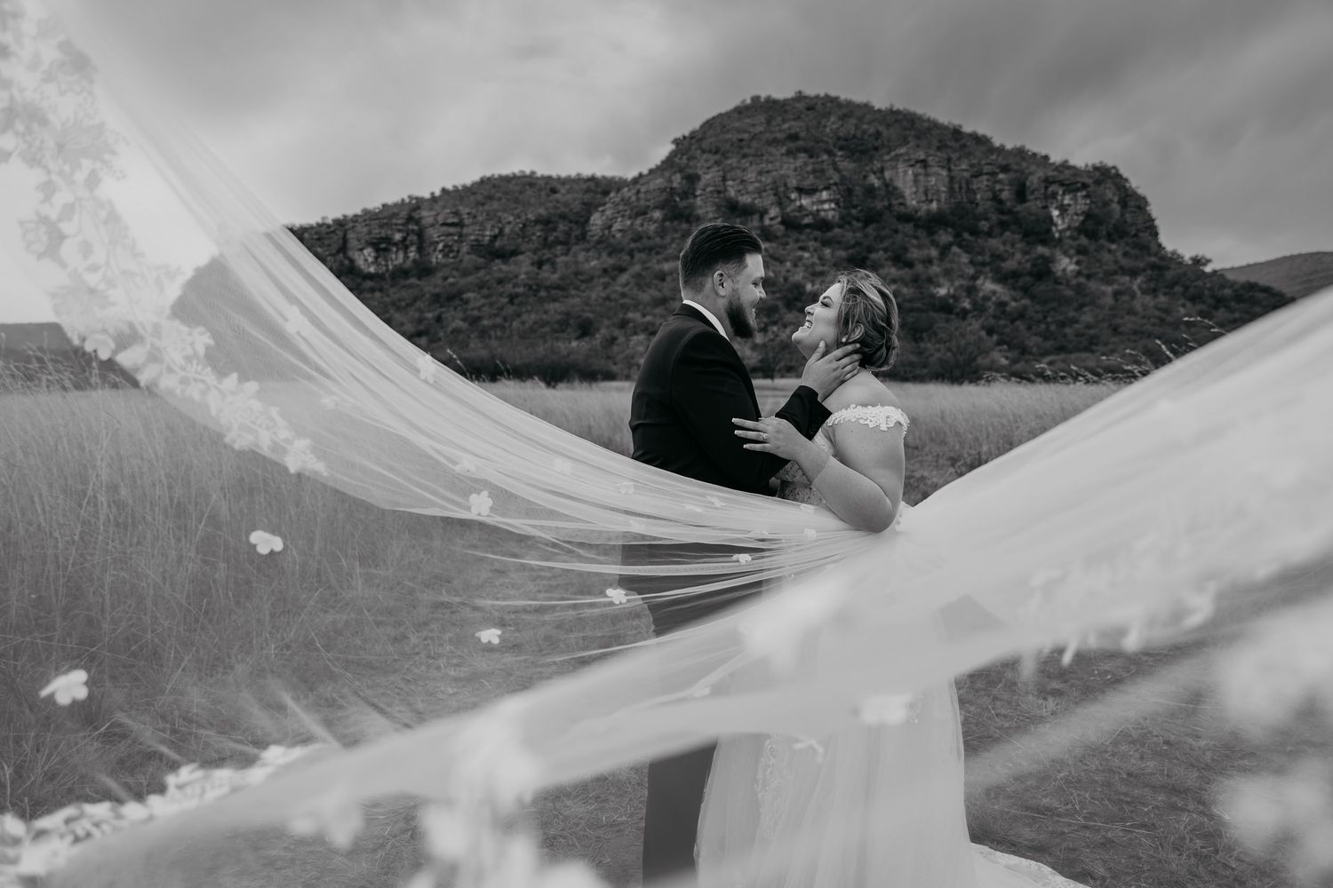 A flowing white veil billows dramatically in the wind against a mountain landscape backdrop.