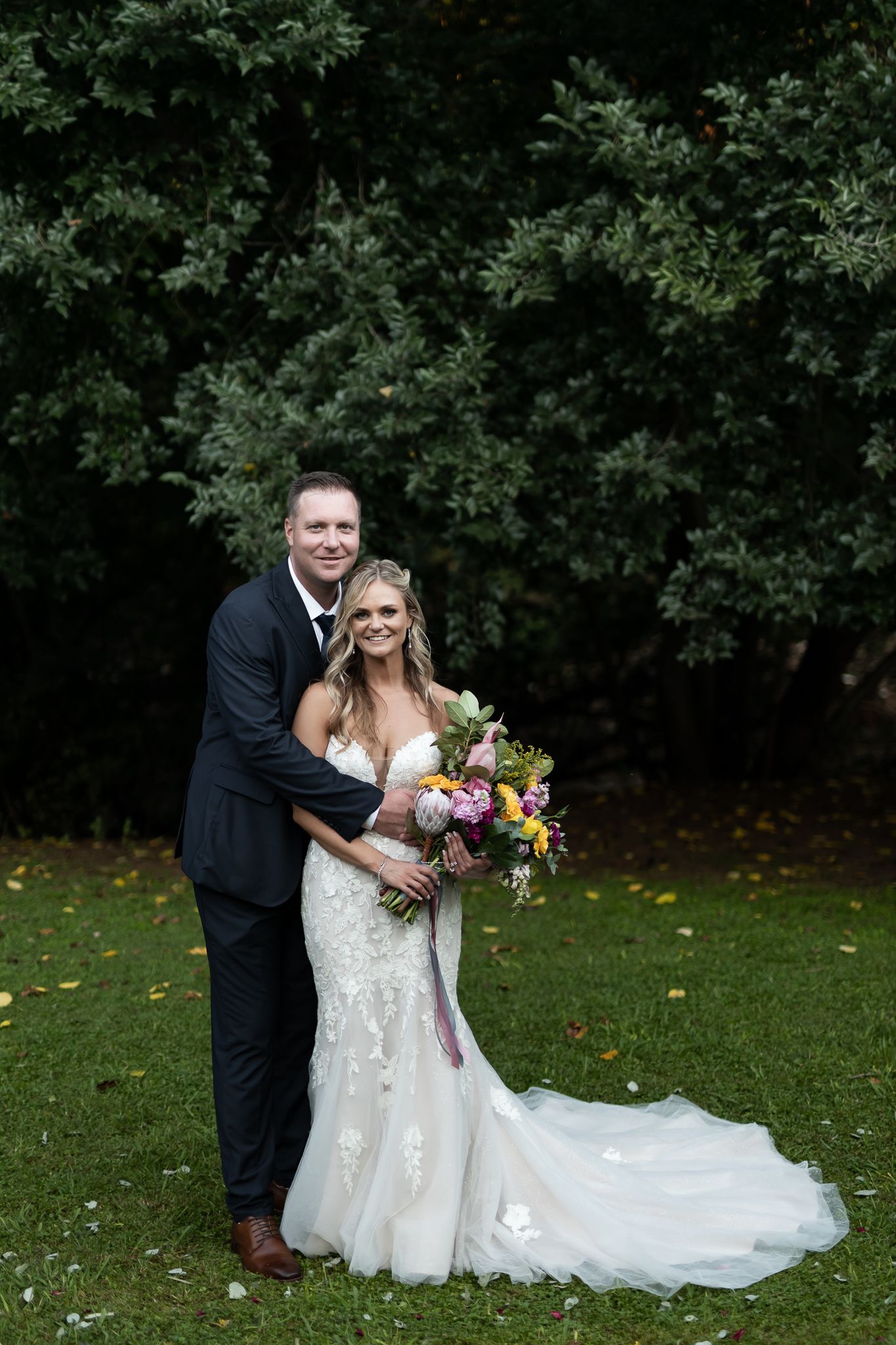 Couple in wedding attire embraces while holding colorful bouquet against green foliage backdrop.
