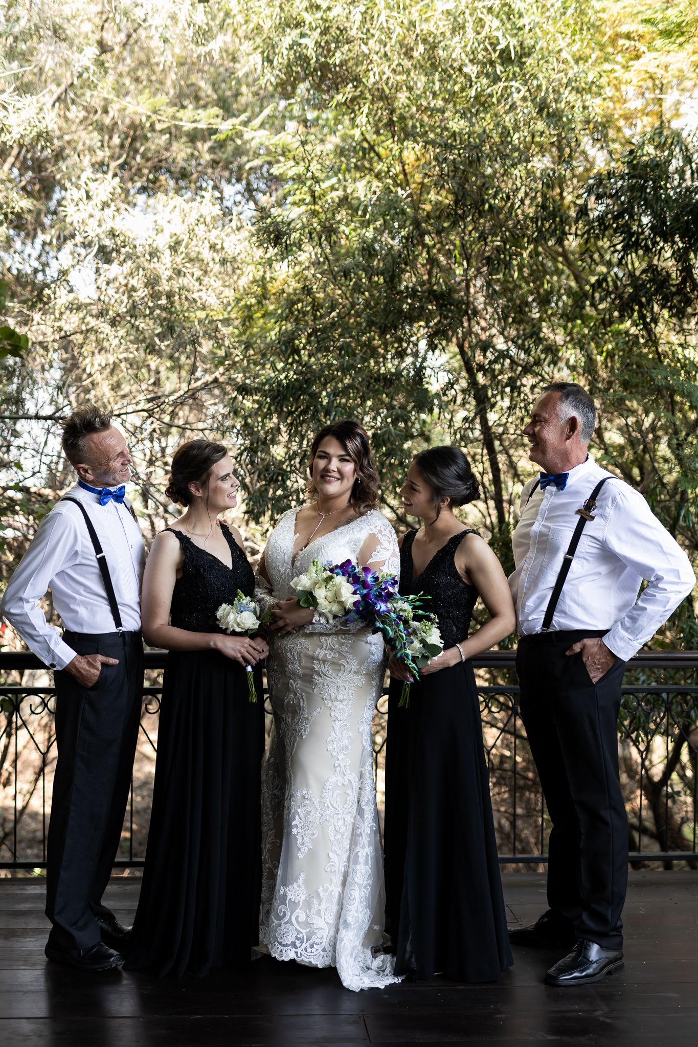 A wedding party poses together in black and white attire on an outdoor deck with greenery in background.