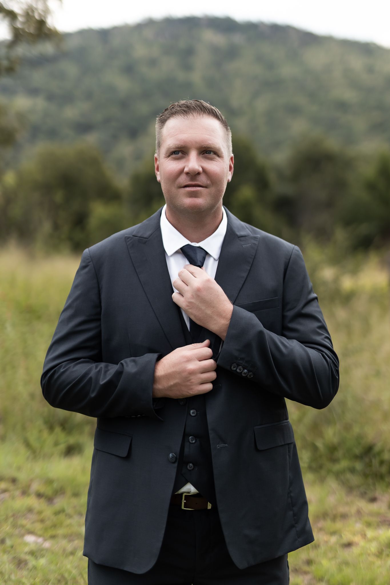 Groom in classic dark suit adjusts tie while posing in outdoor setting with mountain backdrop.