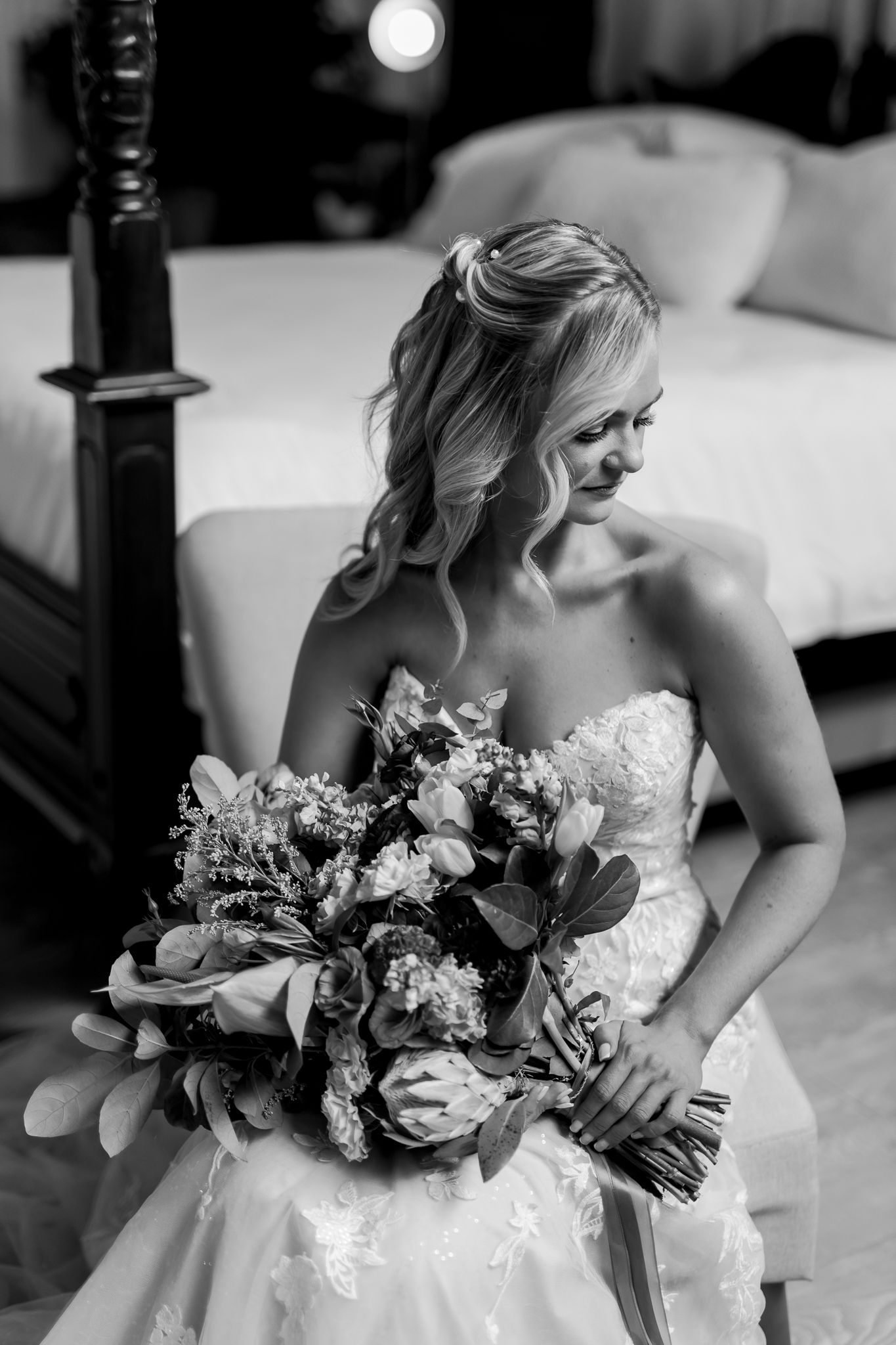 Black and white portrait of bride holding elegant floral bouquet in bedroom setting.