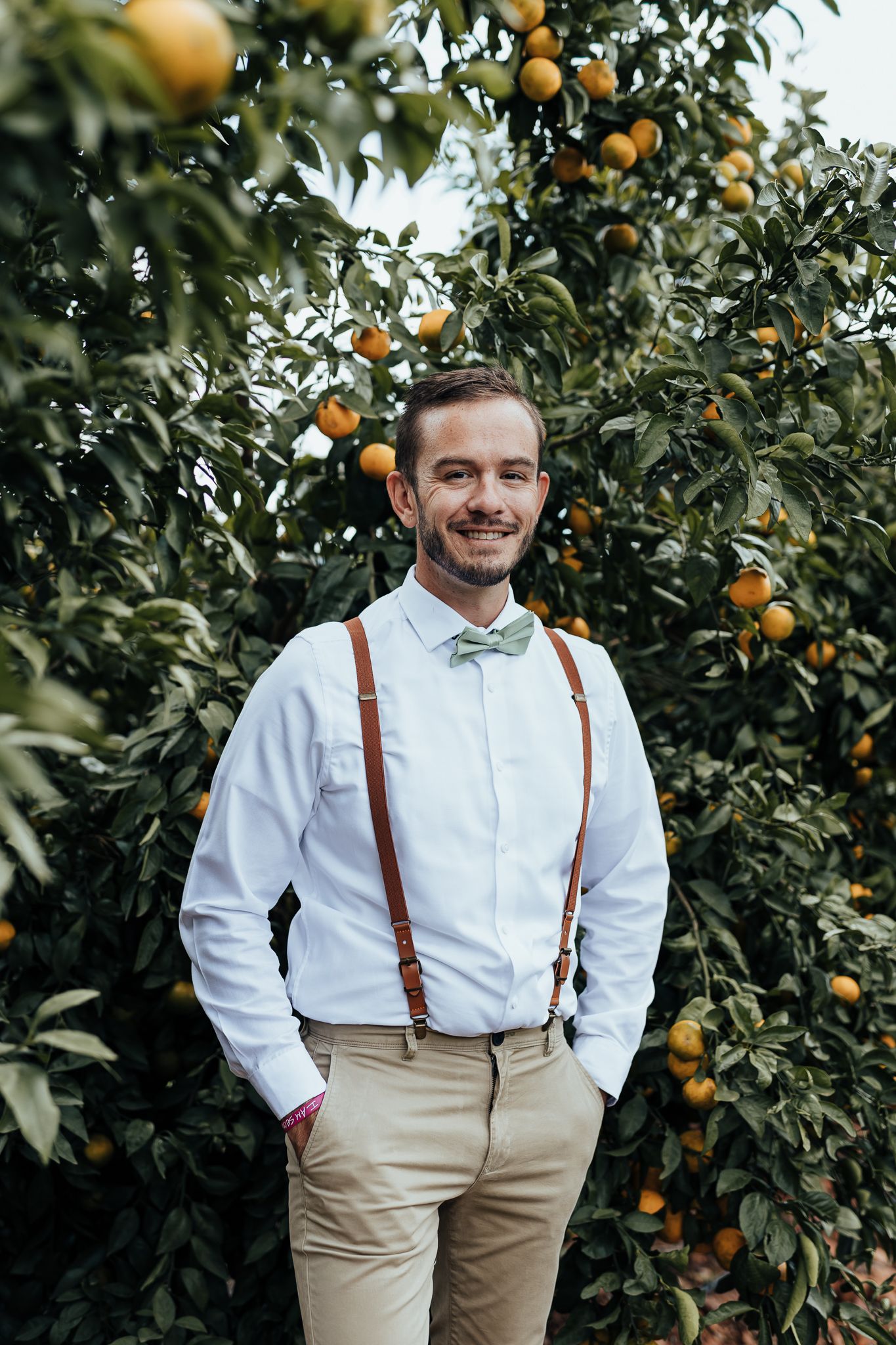 Wedding attendee in white shirt and mint bow tie smiles among citrus trees.