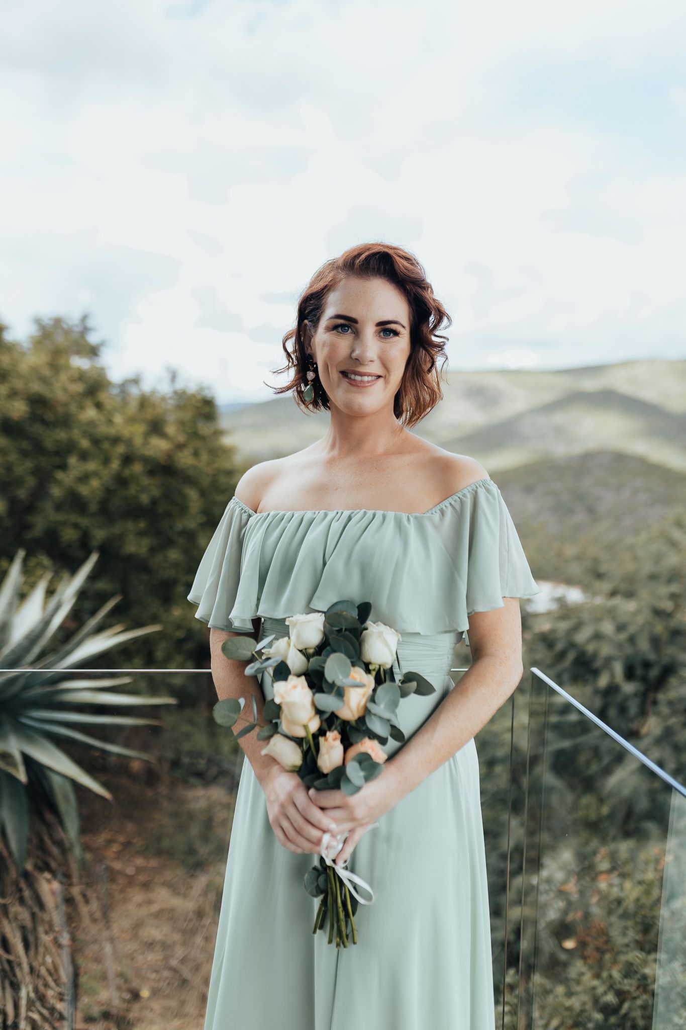 Bridesmaid in sage green off-shoulder dress holds bouquet against mountain landscape.