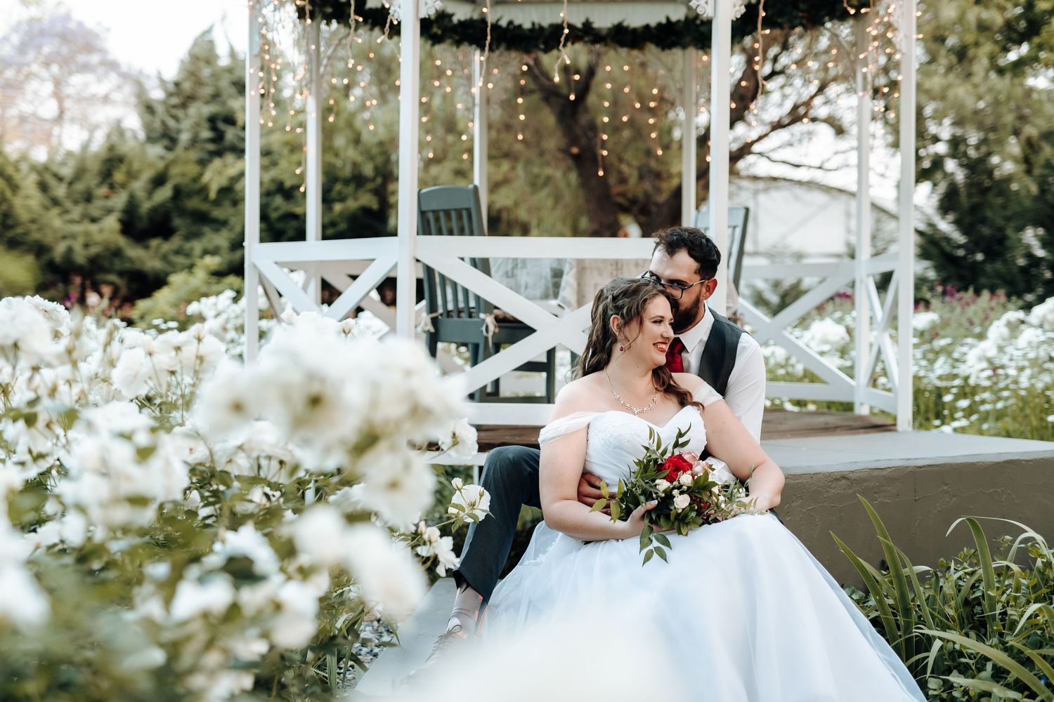 Intimate wedding moment captured in a white gazebo with string lights and flourishing garden.