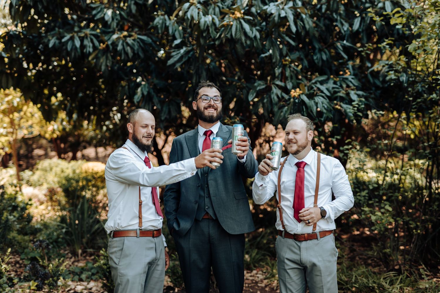 Three groomsmen share drinks and laughter during outdoor wedding photo session.