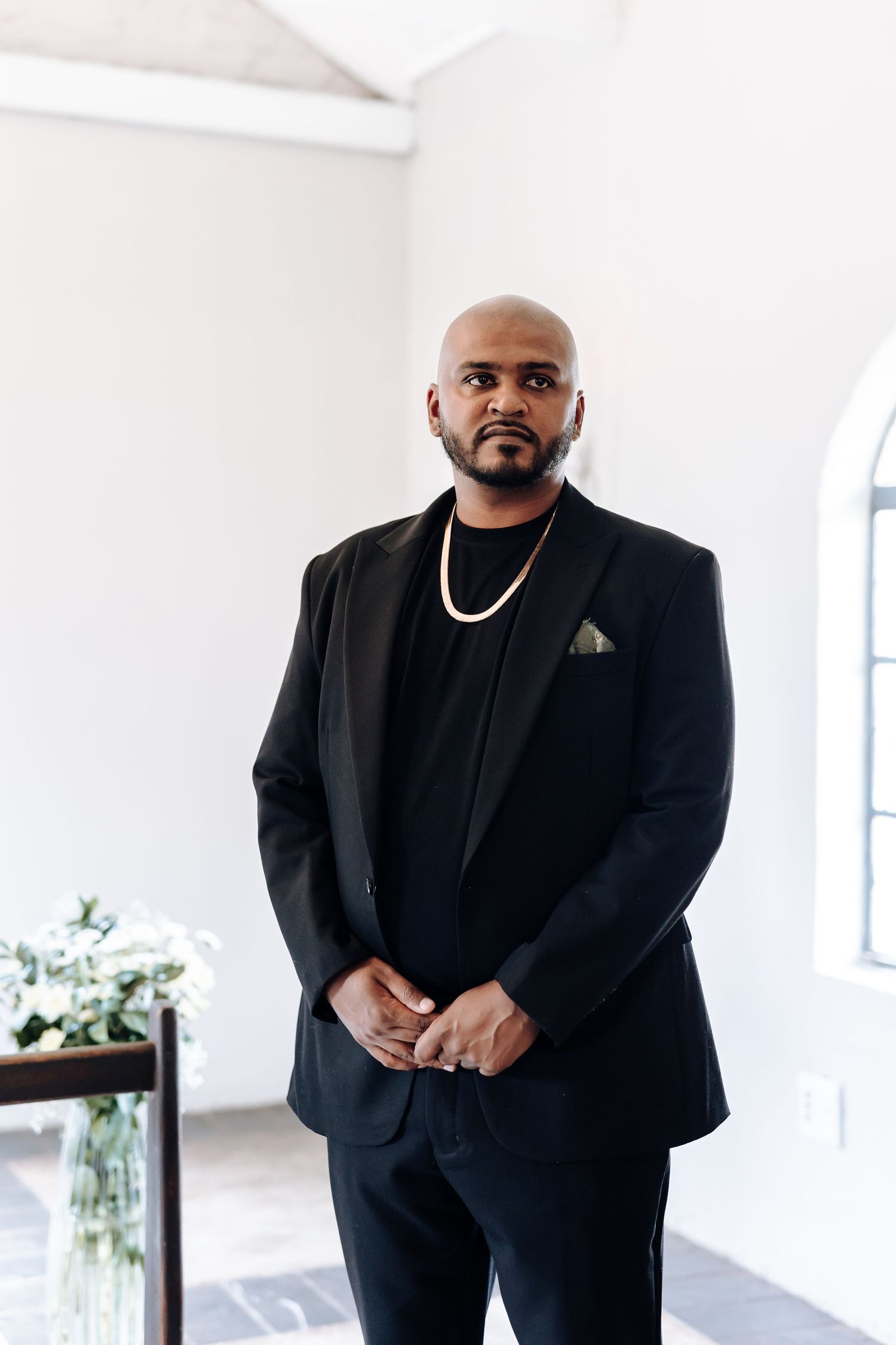 Groom in classic black suit and gold chain stands confidently in white chapel setting.