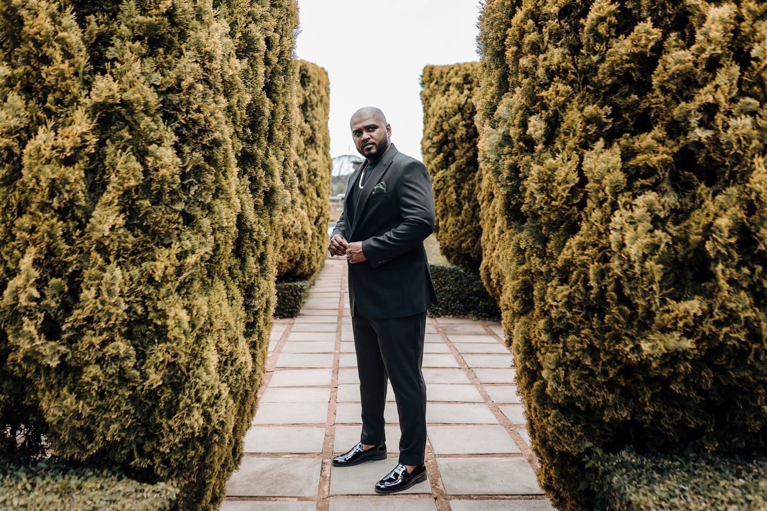 Person in dark suit standing between tall hedges on stone pathway.
