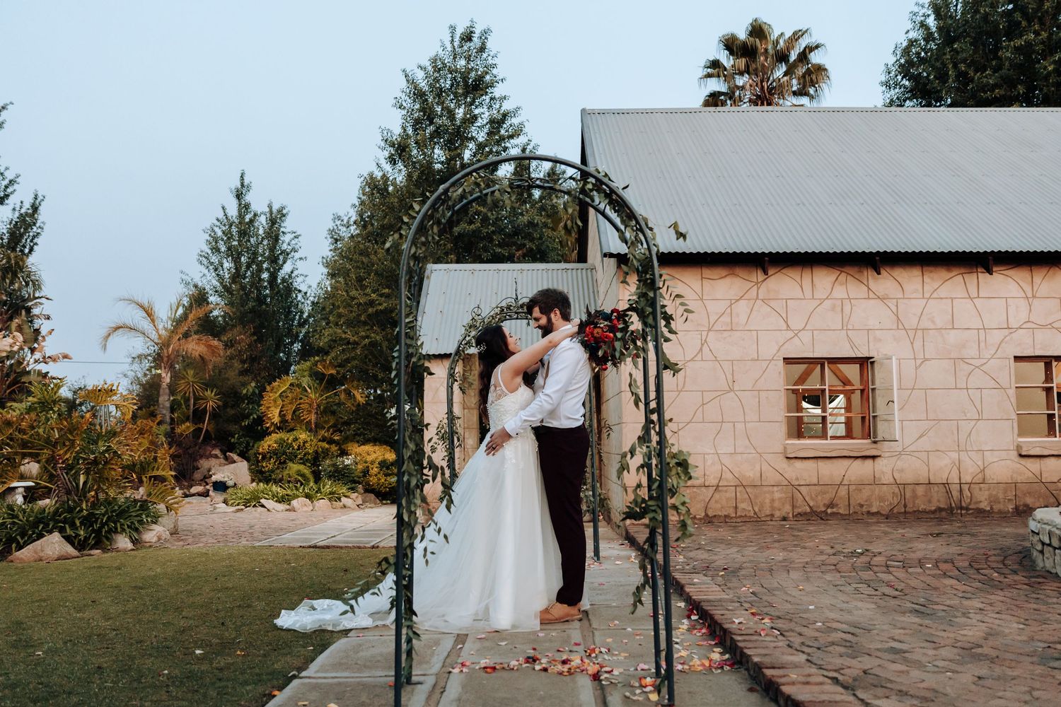 Wedding couple standing under metal arch against rustic stone building exterior.