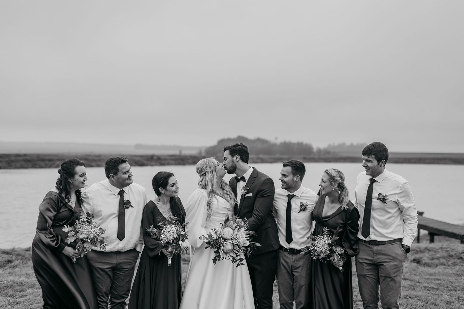 Black and white photo of wedding party laughing together by waterfront dock.