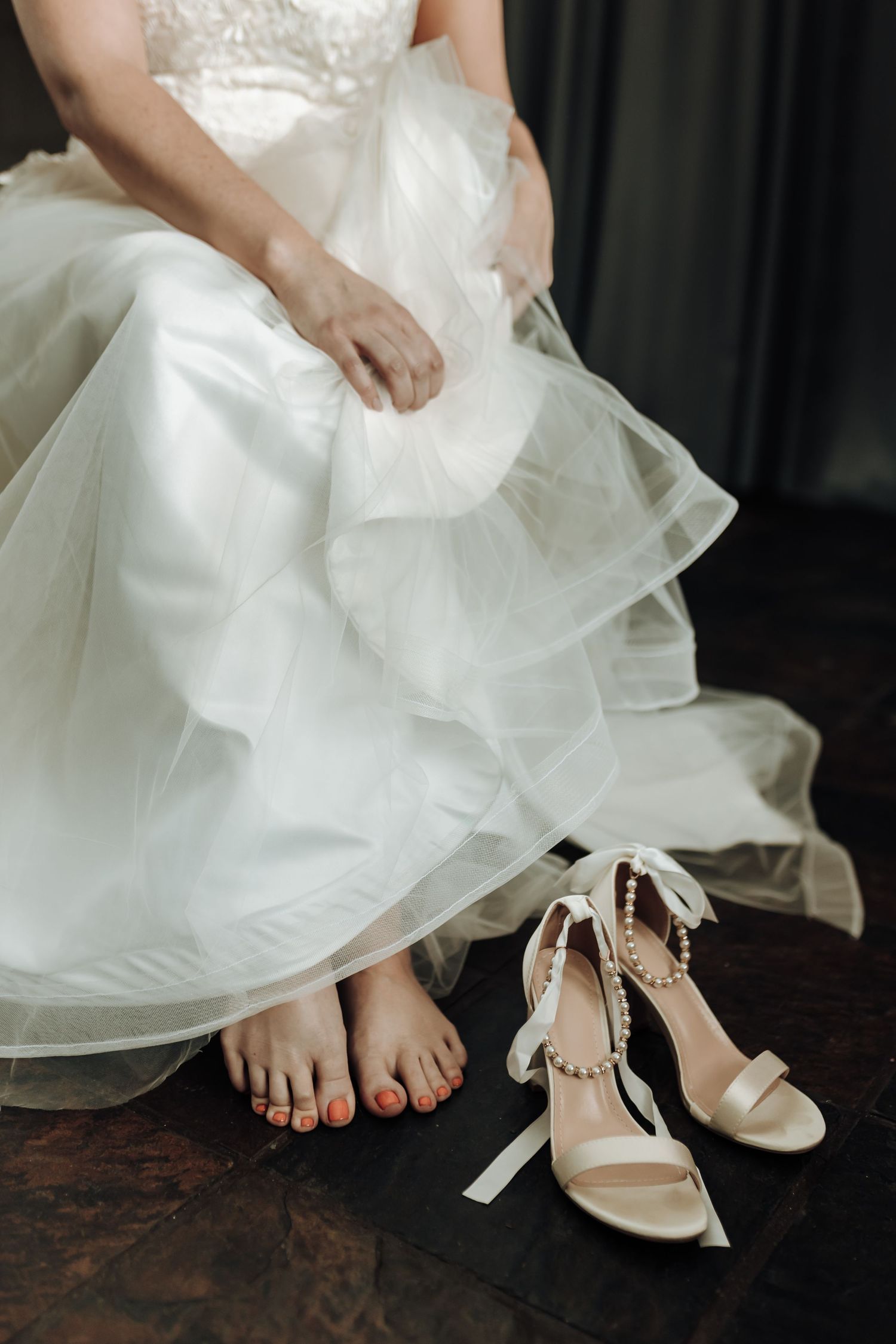 Close-up of wedding dress hem and bridal shoes on dark wooden floor.