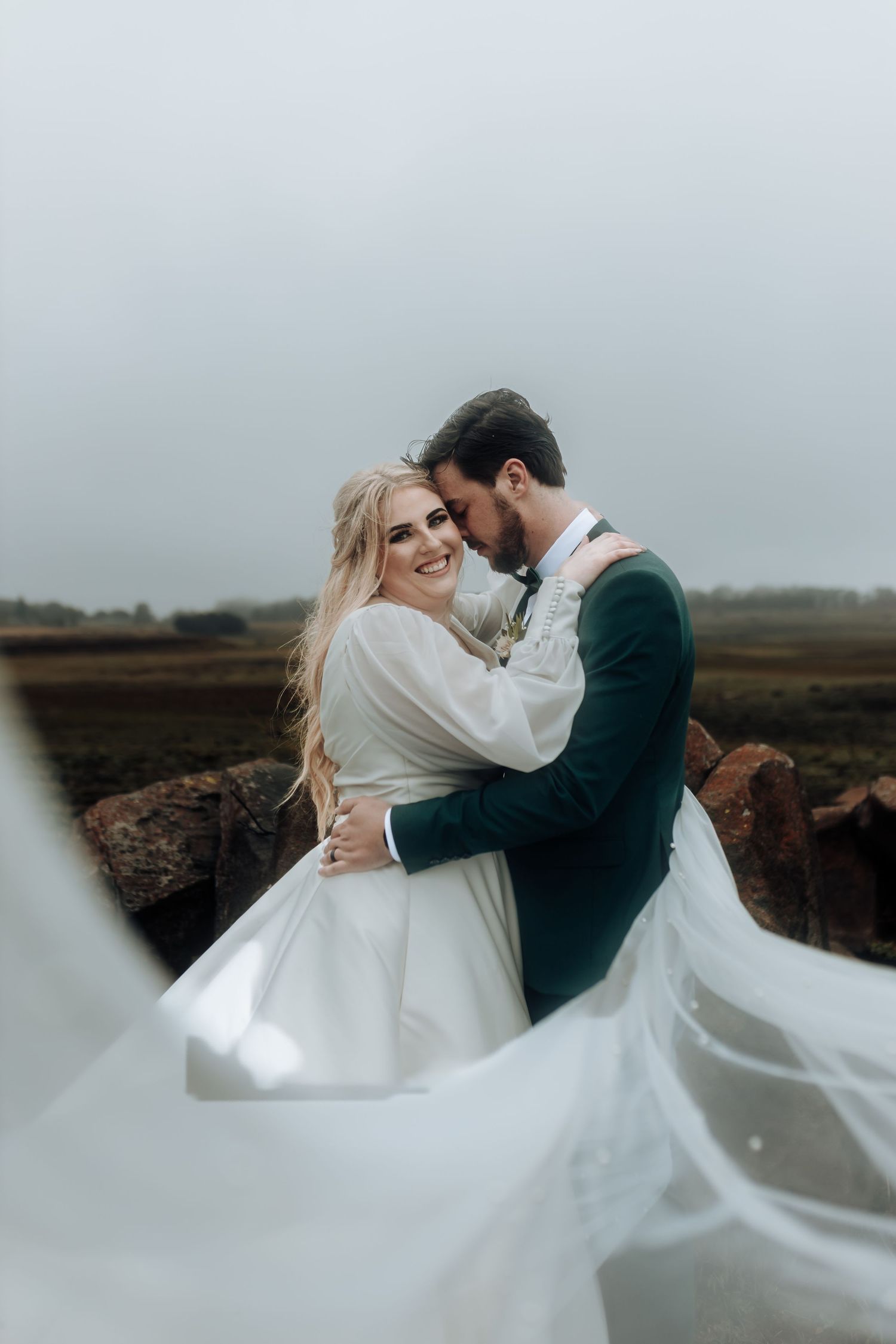 Romantic wedding couple embraces on a misty hilltop as the bride's dress flows dramatically in the wind.
