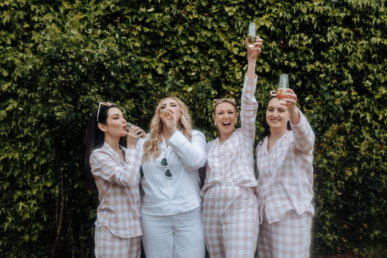 Four friends in matching pajamas celebrate joyfully against a green hedge backdrop during a special occasion.