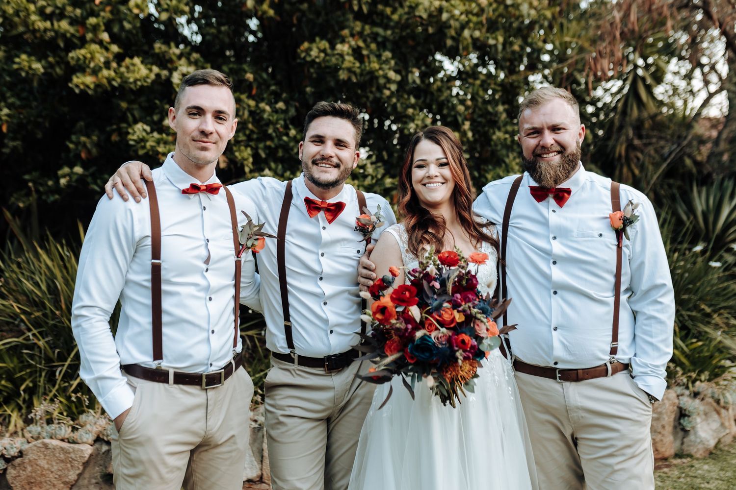 Wedding party members wearing light blue shirts with suspenders and bow ties pose with bride holding vibrant red bouquet.