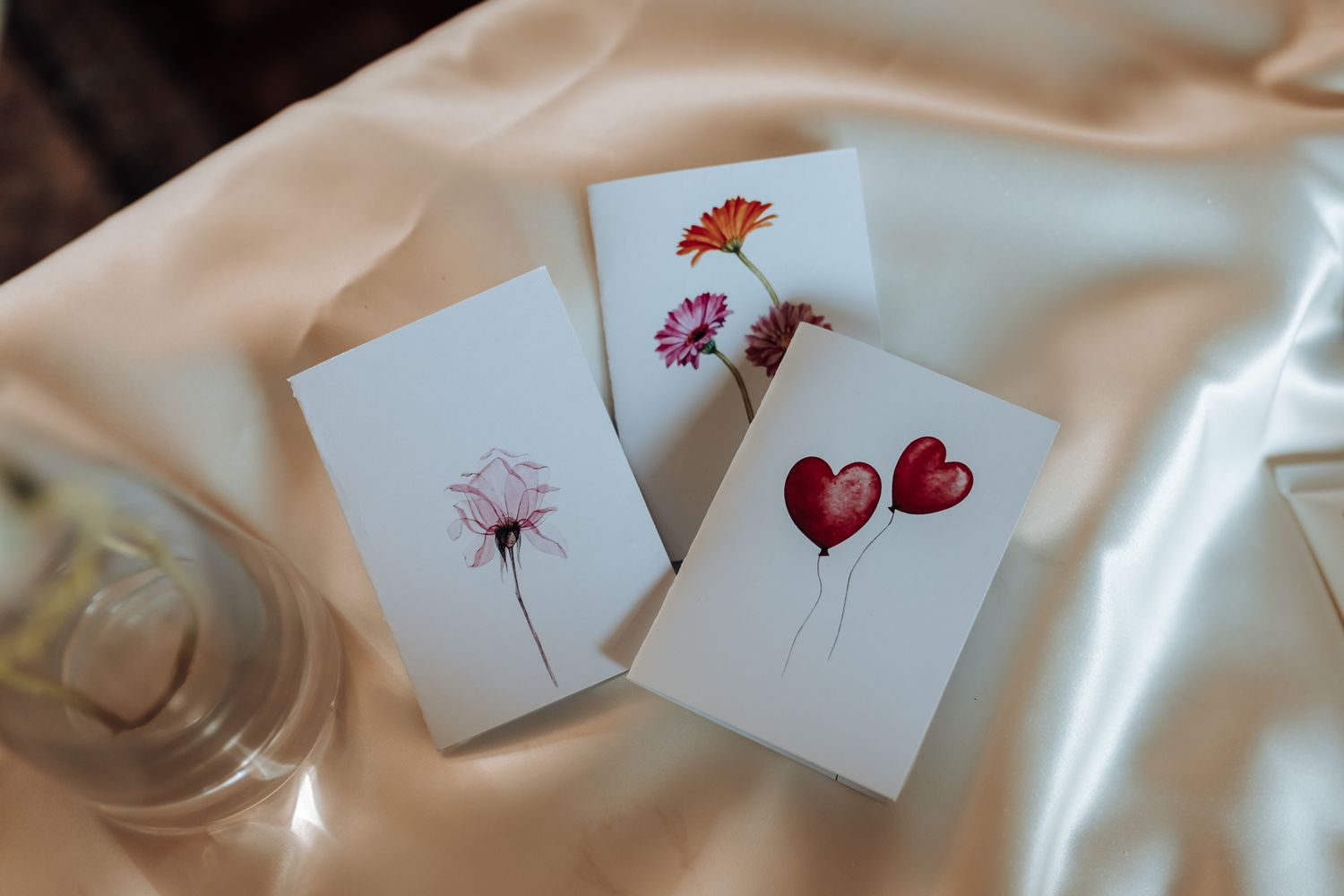 Three delicate watercolor cards featuring floral designs and red heart illustrations lay on silky fabric.