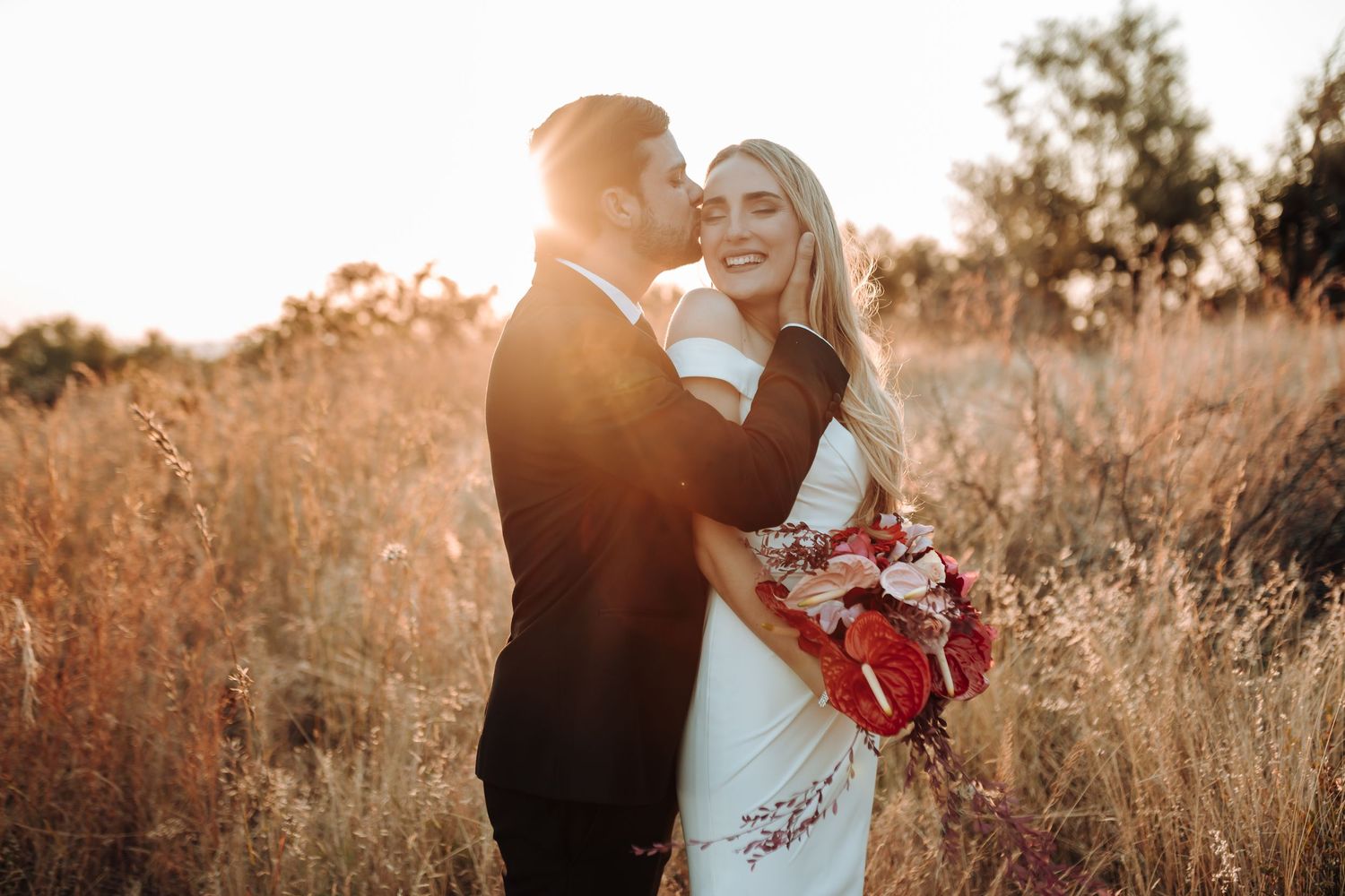 A romantic wedding couple embraces in golden sunlight amidst tall grass at sunset with a red bouquet.