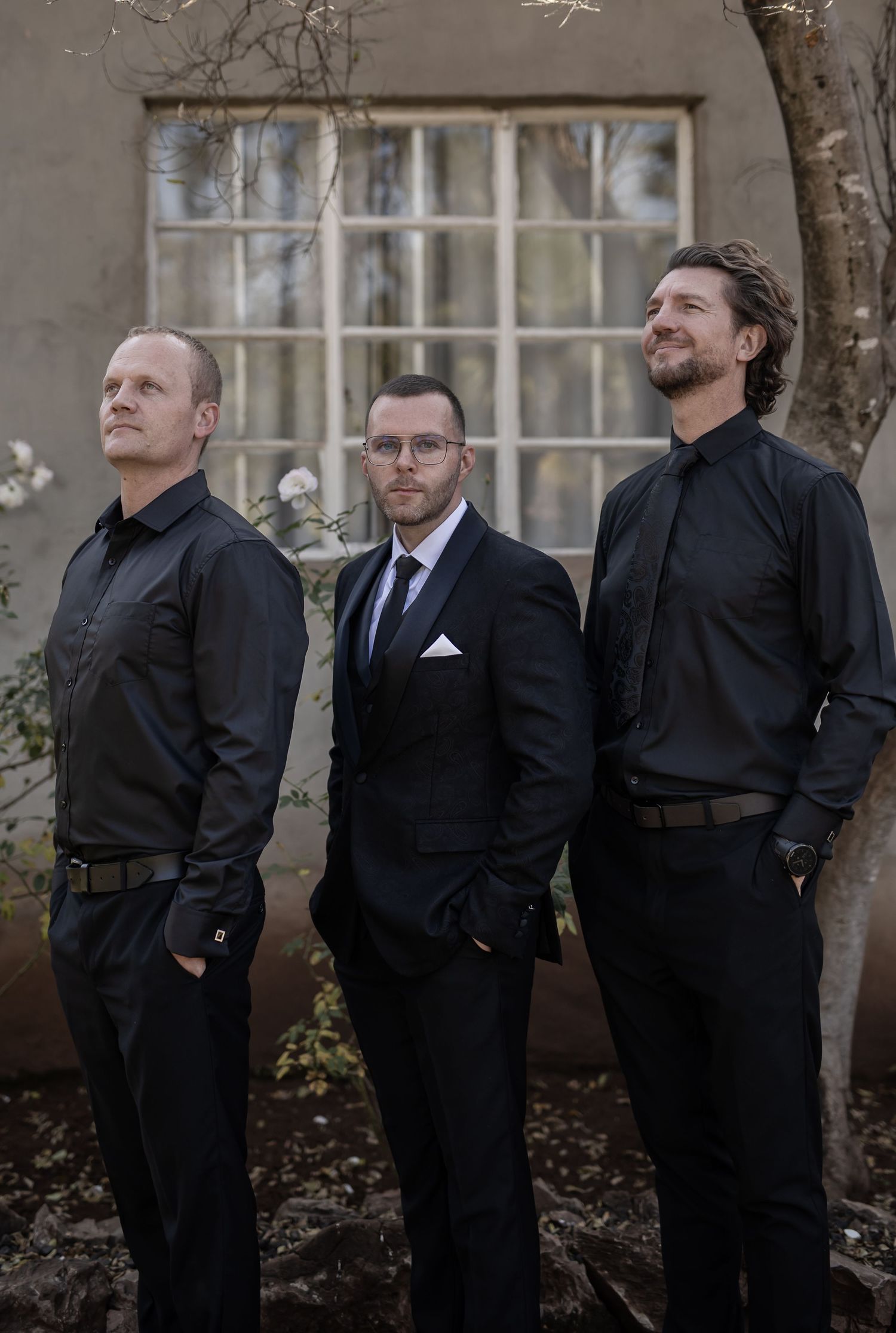 Three men in dark formal attire pose outdoors against a gray building with a white window frame in the background.