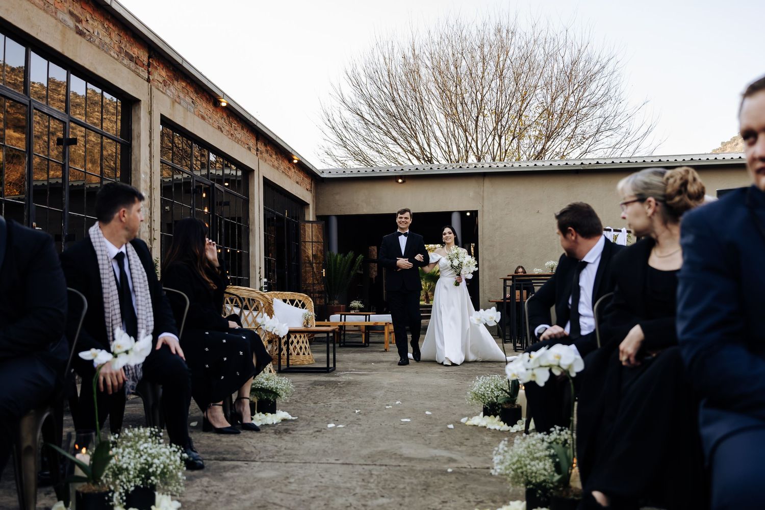 Outdoor wedding ceremony at rustic venue with simple floral decor and guests gathered on patio.
