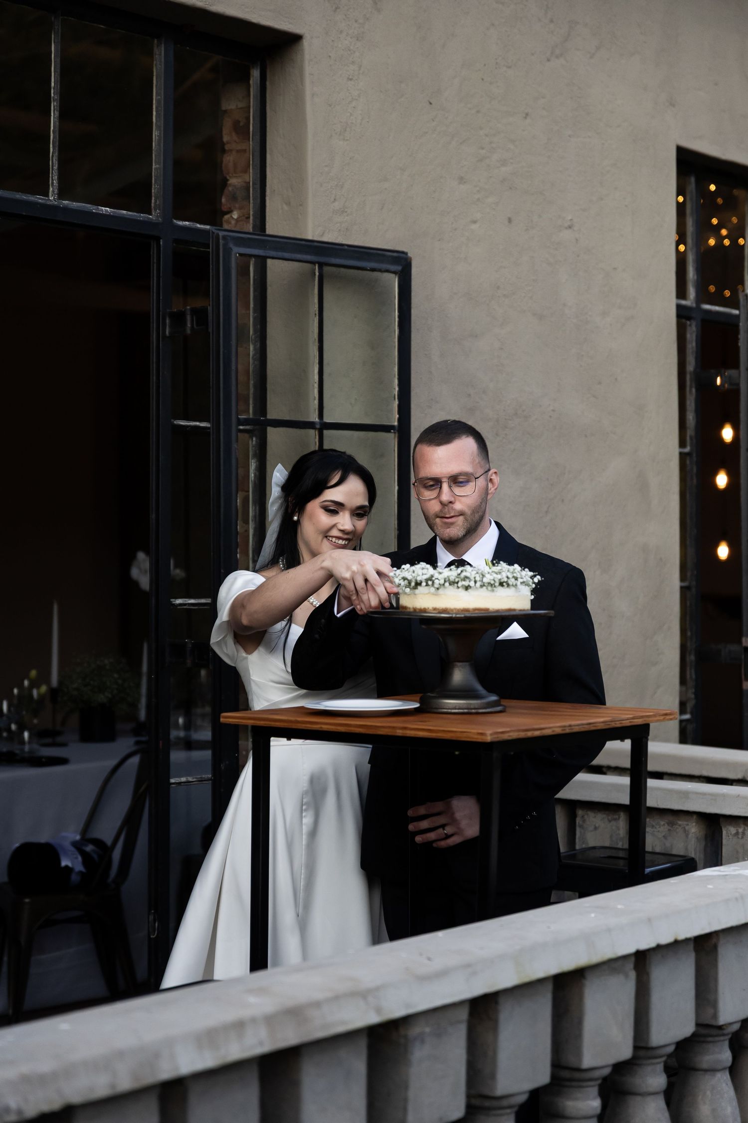 Couple in formal wedding attire stands on balcony cutting into a white cake displayed on wooden table.