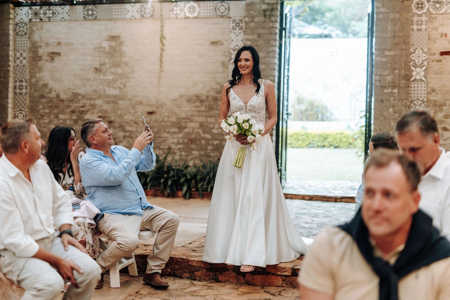 Bride in white gown holding bouquet smiles at guests during wedding ceremony in rustic brick venue.