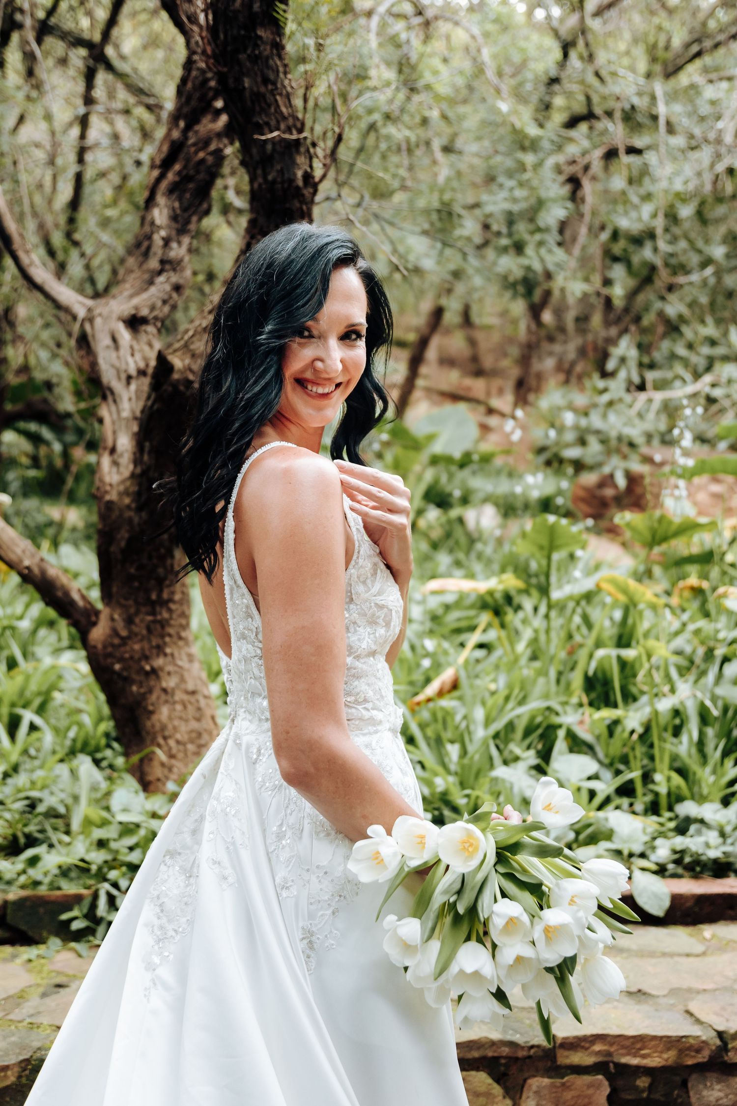 Smiling bride in white lace gown holds white tulip bouquet in lush garden setting.