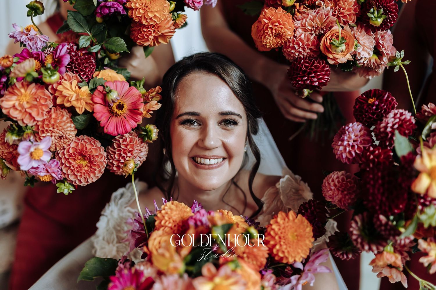 Smiling bride surrounded by vibrant orange and pink dahlia bouquets held by bridesmaids on her wedding day.
