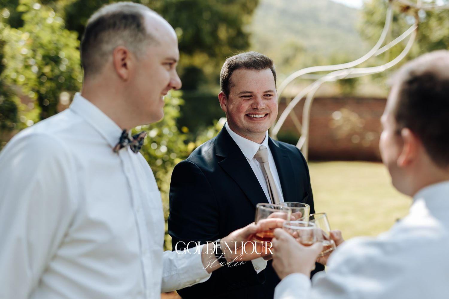 Three groomsmen in suits toasting with glasses at an outdoor wedding celebration.