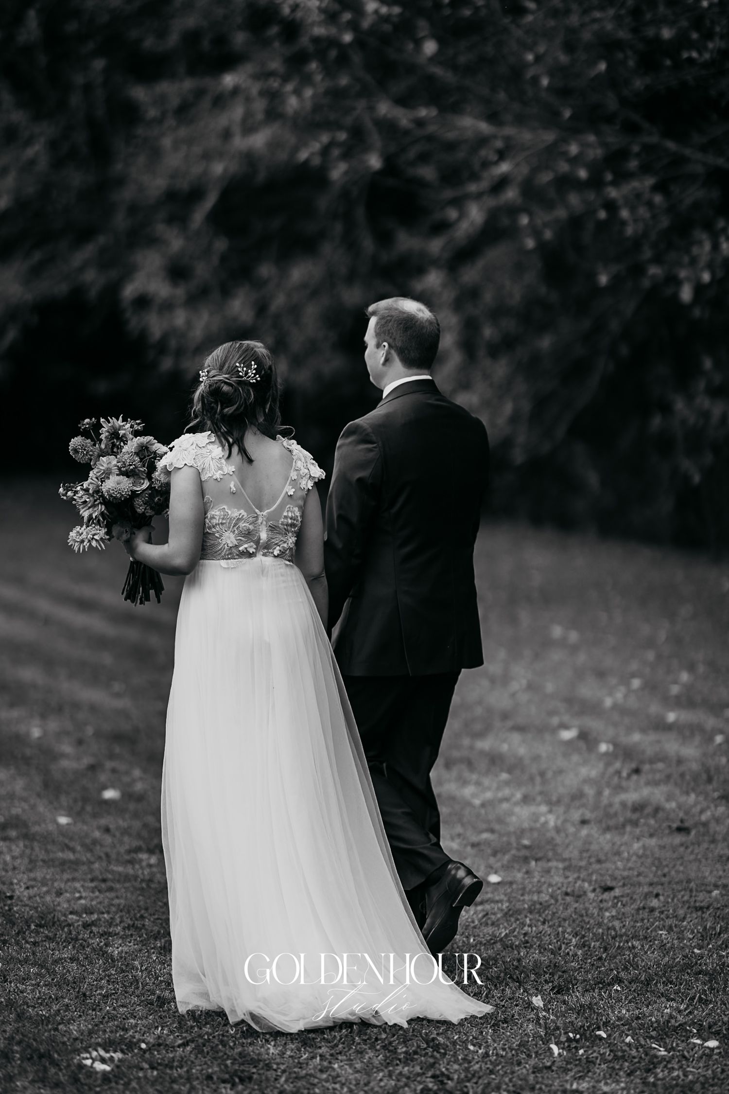 Black and white photo of bride and groom walking hand-in-hand through lush garden.