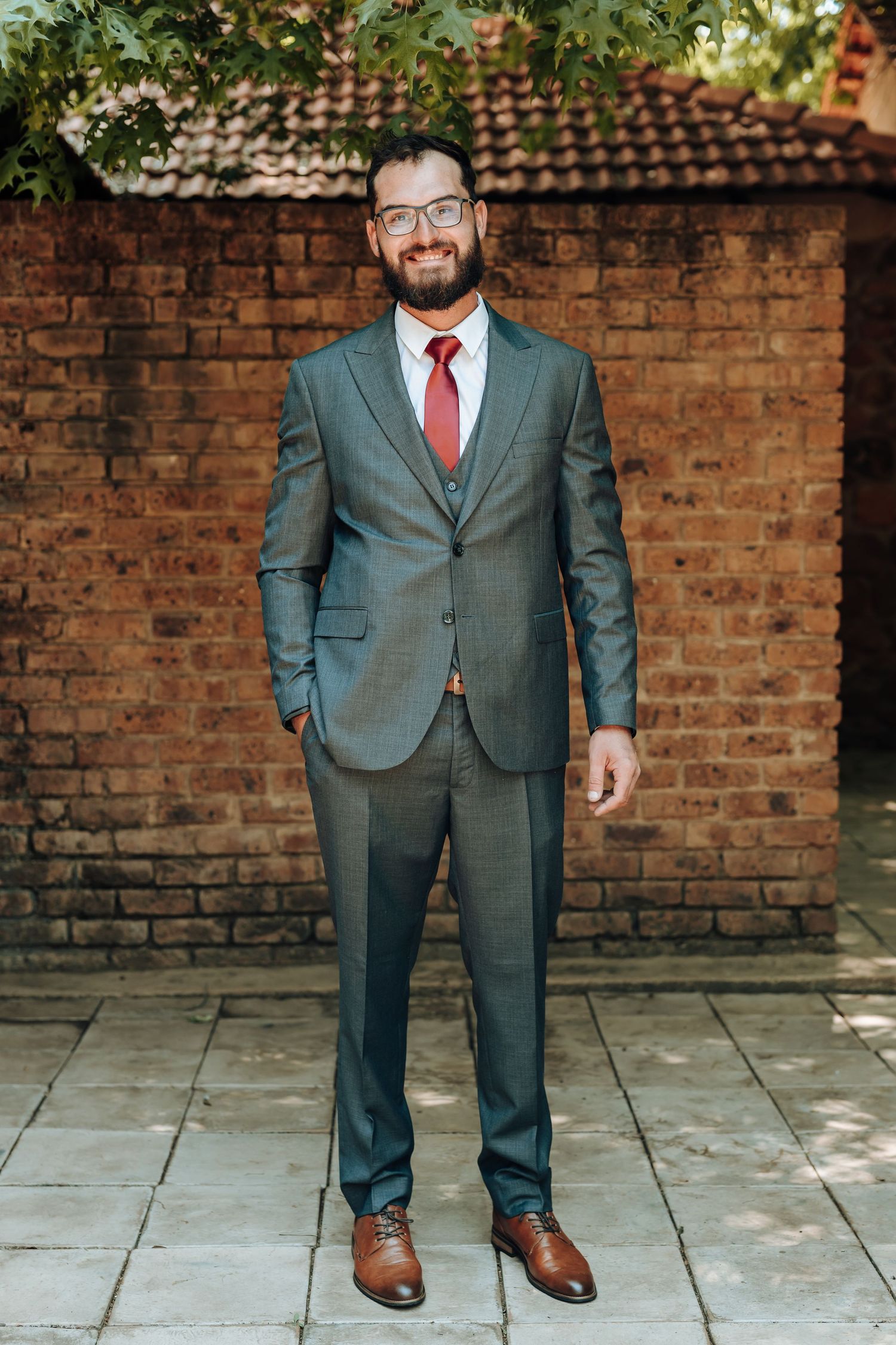 Full length portrait in tailored grey suit against brick wall with overhanging greenery.