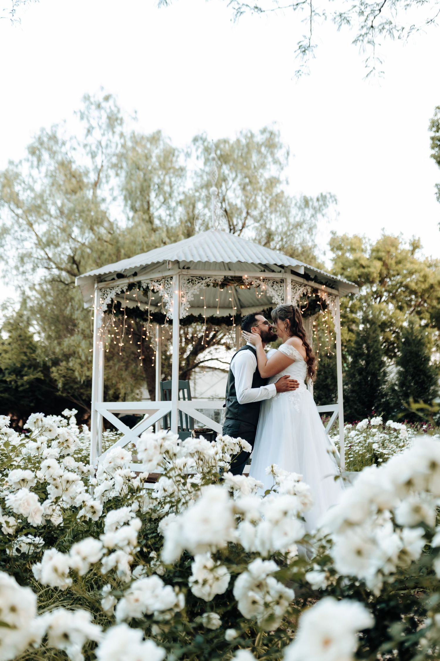 Couple sharing an intimate moment in an ornate white gazebo surrounded by white flower gardens.