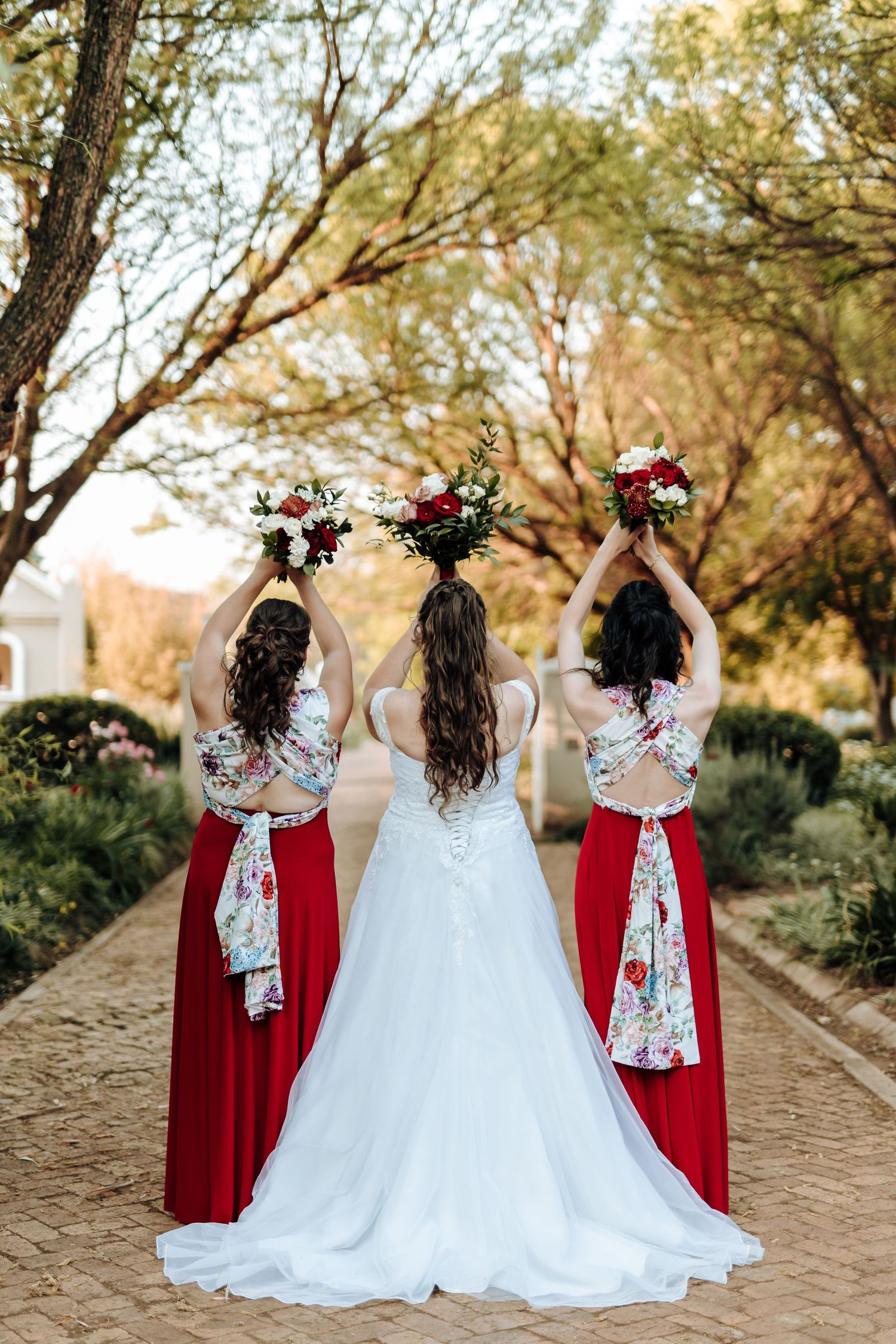 Three women in red dresses raise their bouquets while facing away on a brick pathway.
