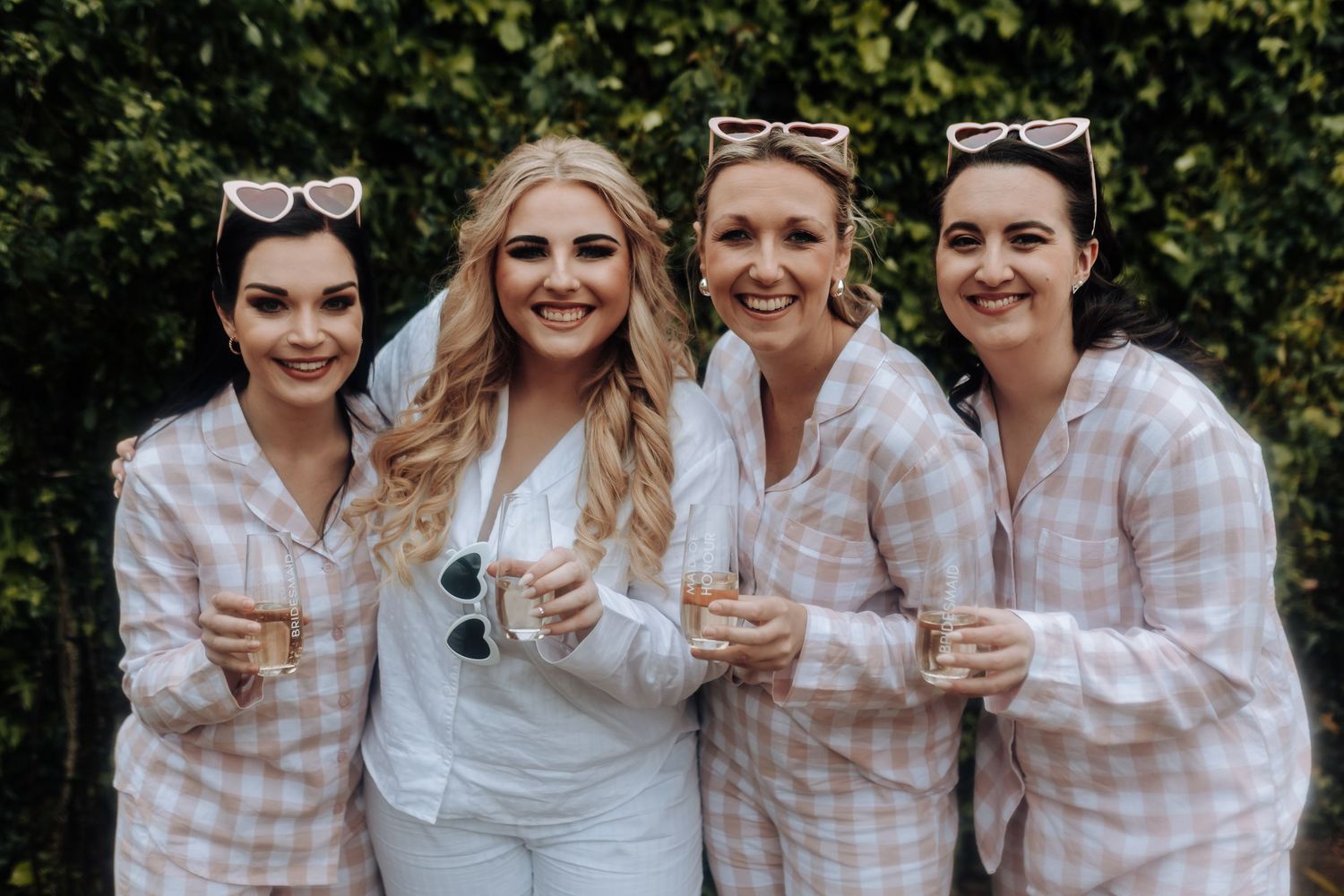 A group in matching pink checkered pajamas poses with champagne glasses against greenery.