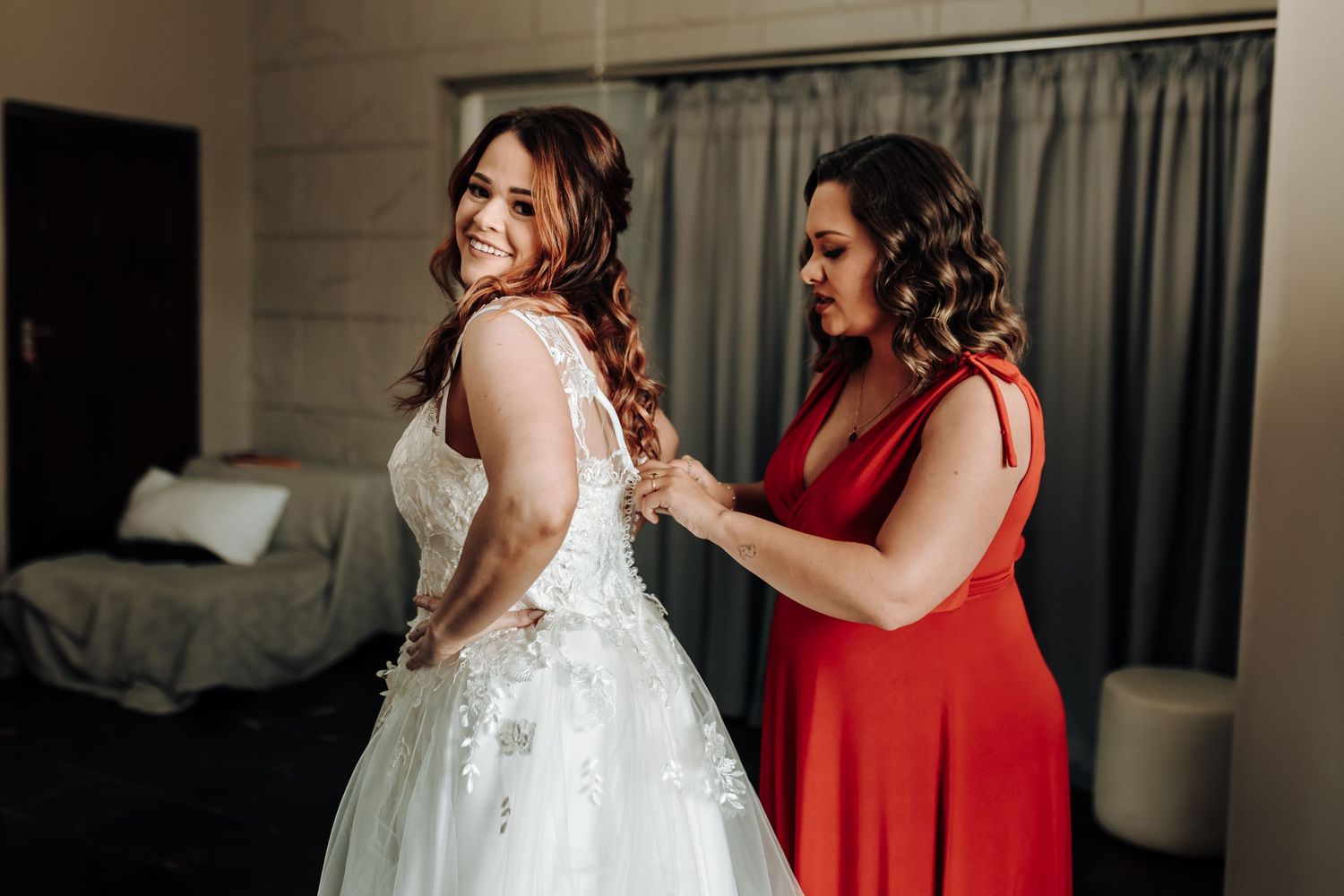 A bride in a white lace gown gets help from a bridesmaid in a red dress while getting ready for the wedding.