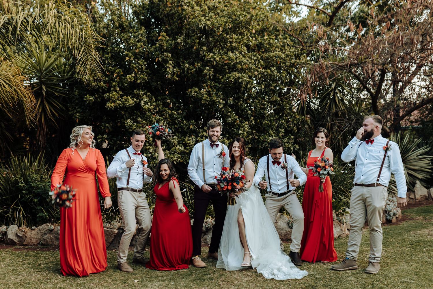 Wedding party poses casually in garden setting wearing red dresses and khaki pants with suspenders.