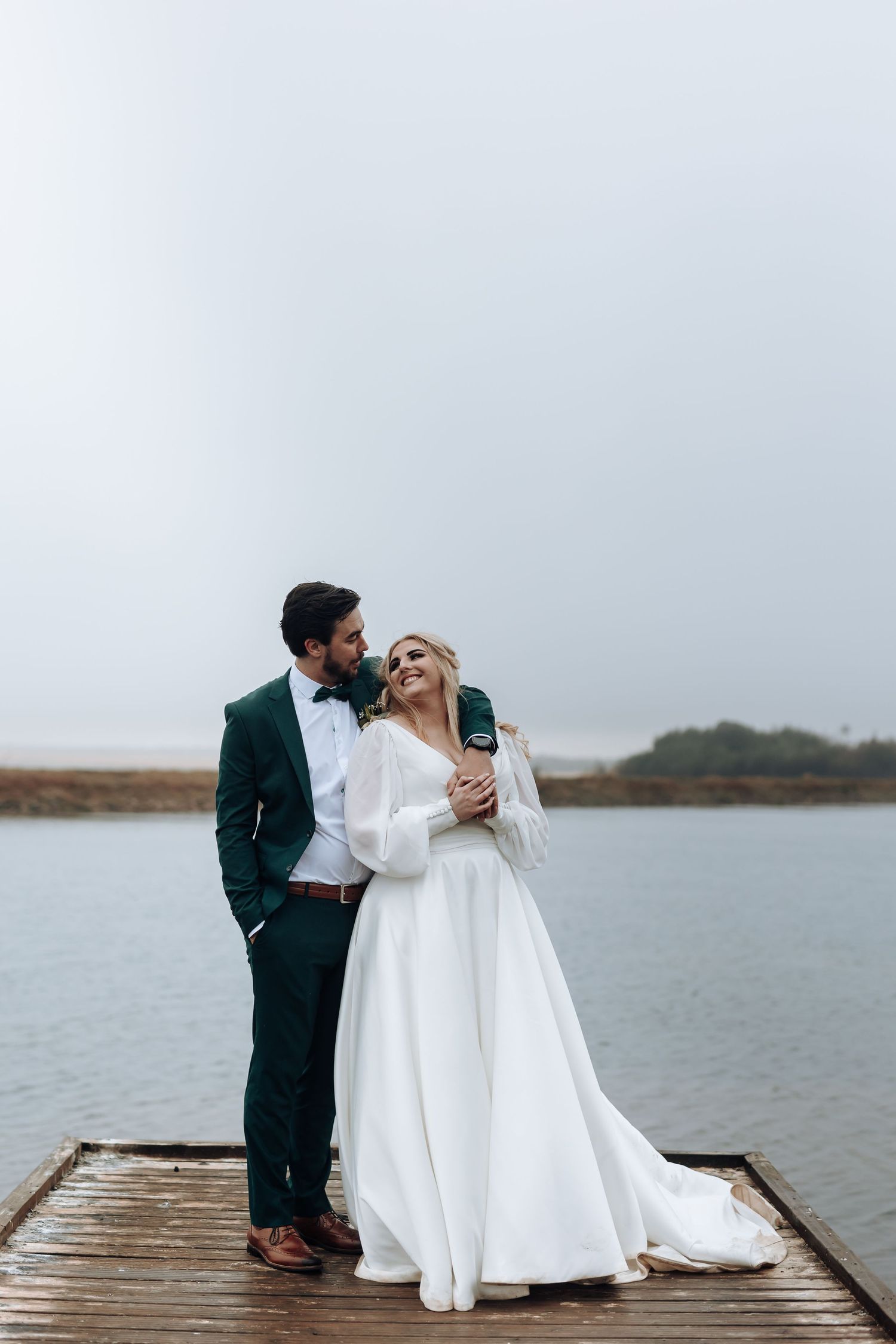Elegant wedding couple stands together on wooden dock with emerald suit and flowing white dress.