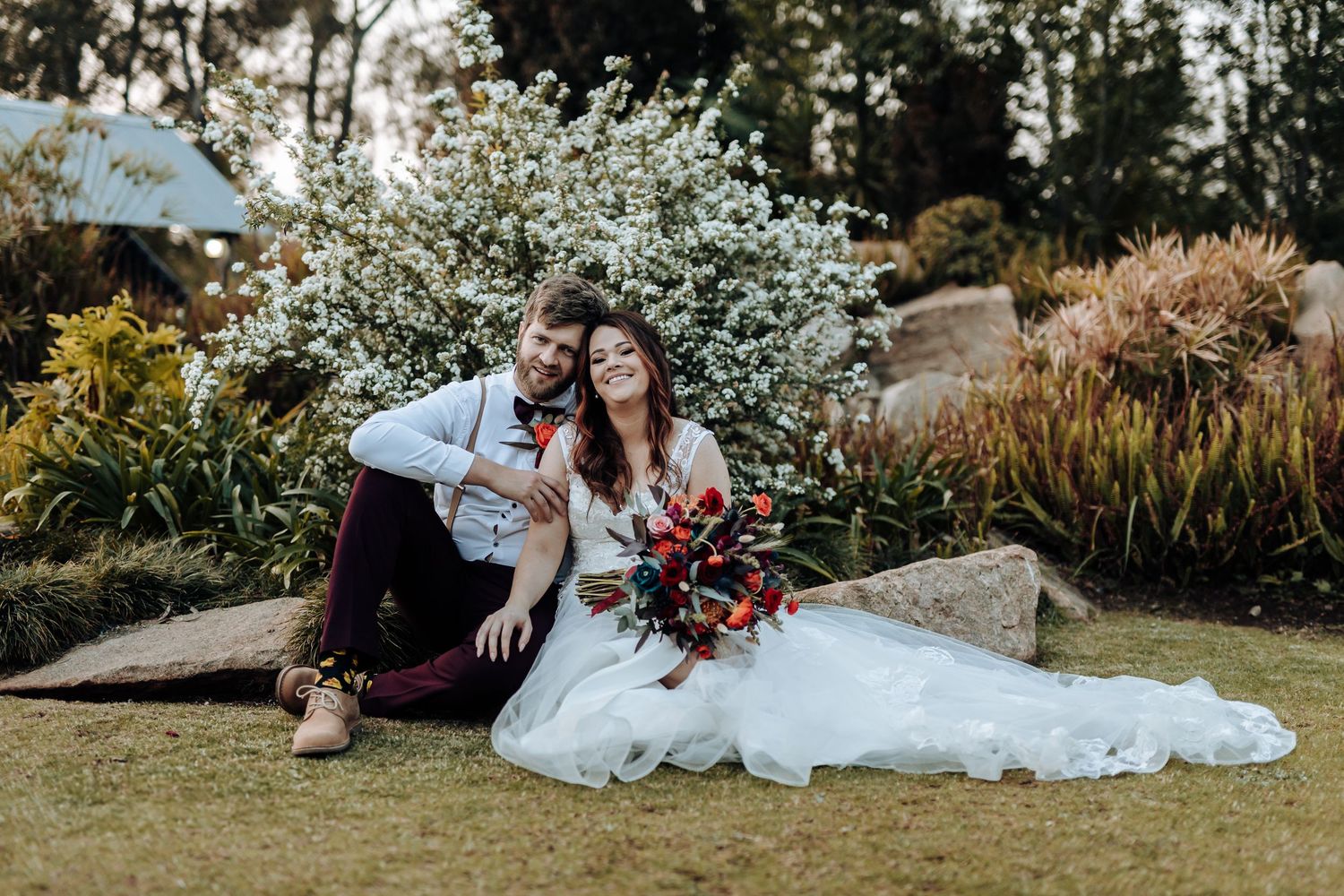Wedding couple sitting on rocks in a garden with blooming white flowers and natural landscaping in background.