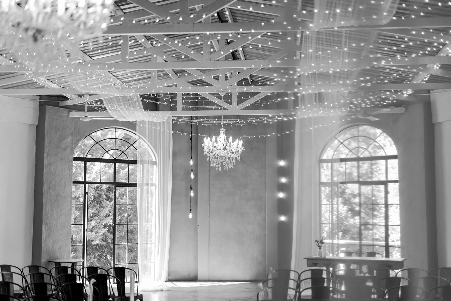 A black and white photo of an elegant wedding venue with string lights and large arched windows.