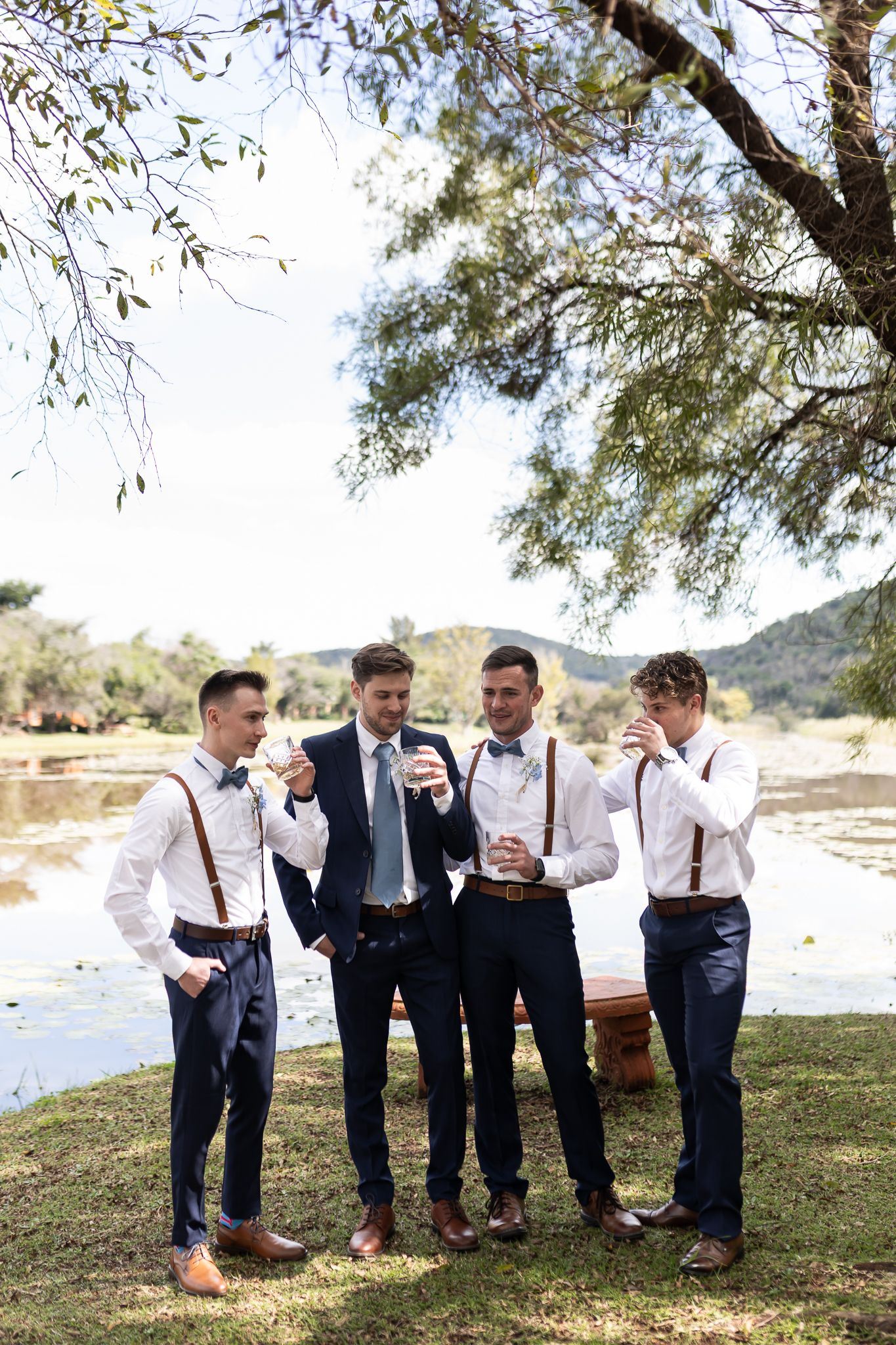 Wedding party members sharing drinks and laughs under tree by lake.