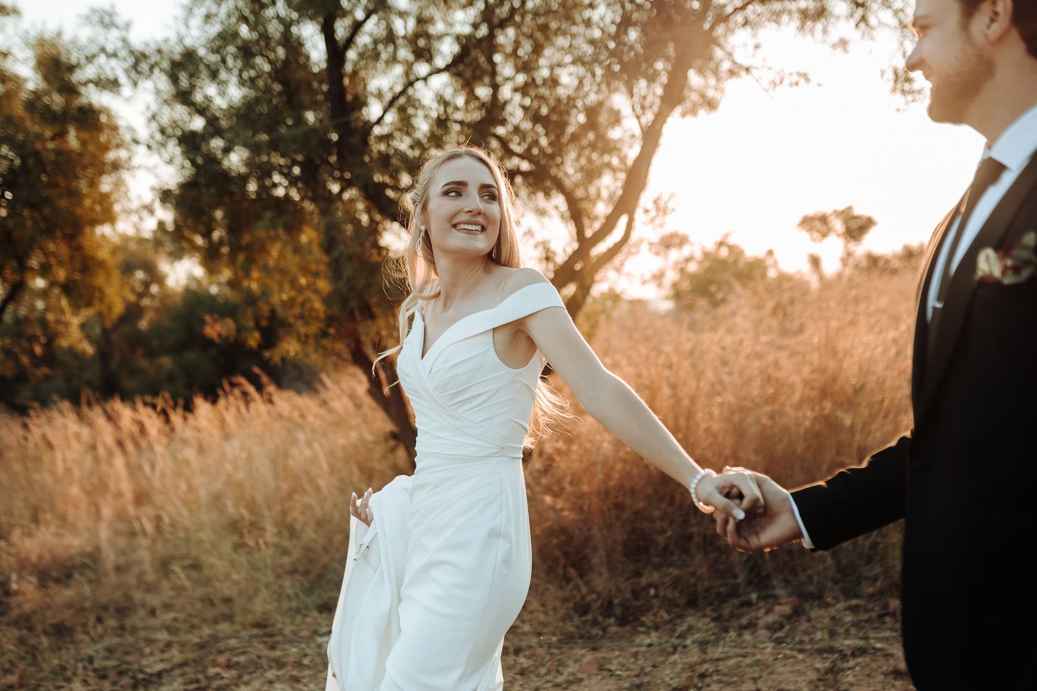 Joyful bride in fitted white gown leads groom through golden field during sunset wedding photoshoot.