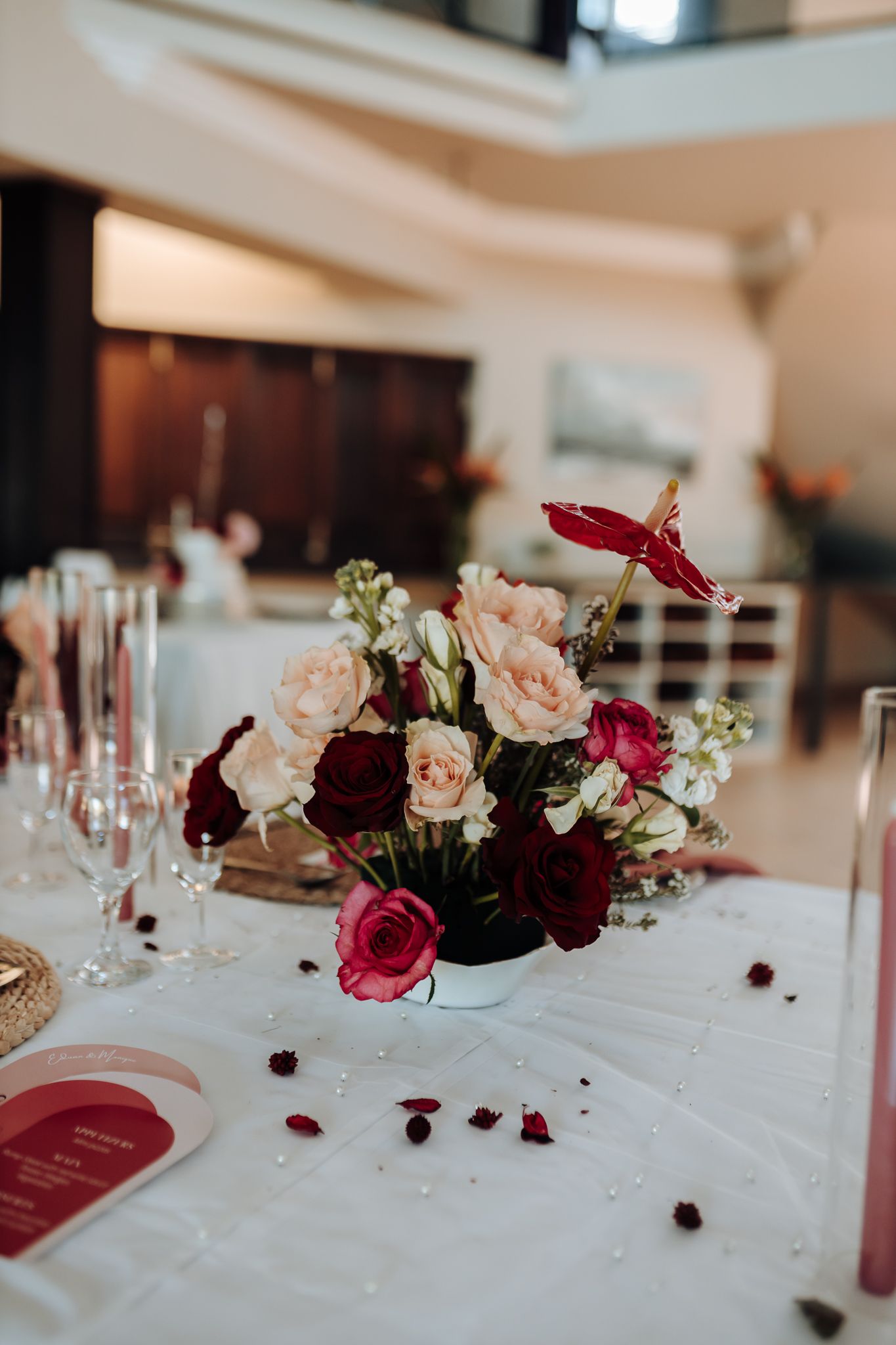 Elegant floral centerpiece with pink and burgundy roses on a white tablecloth with scattered petals and wine glasses.
