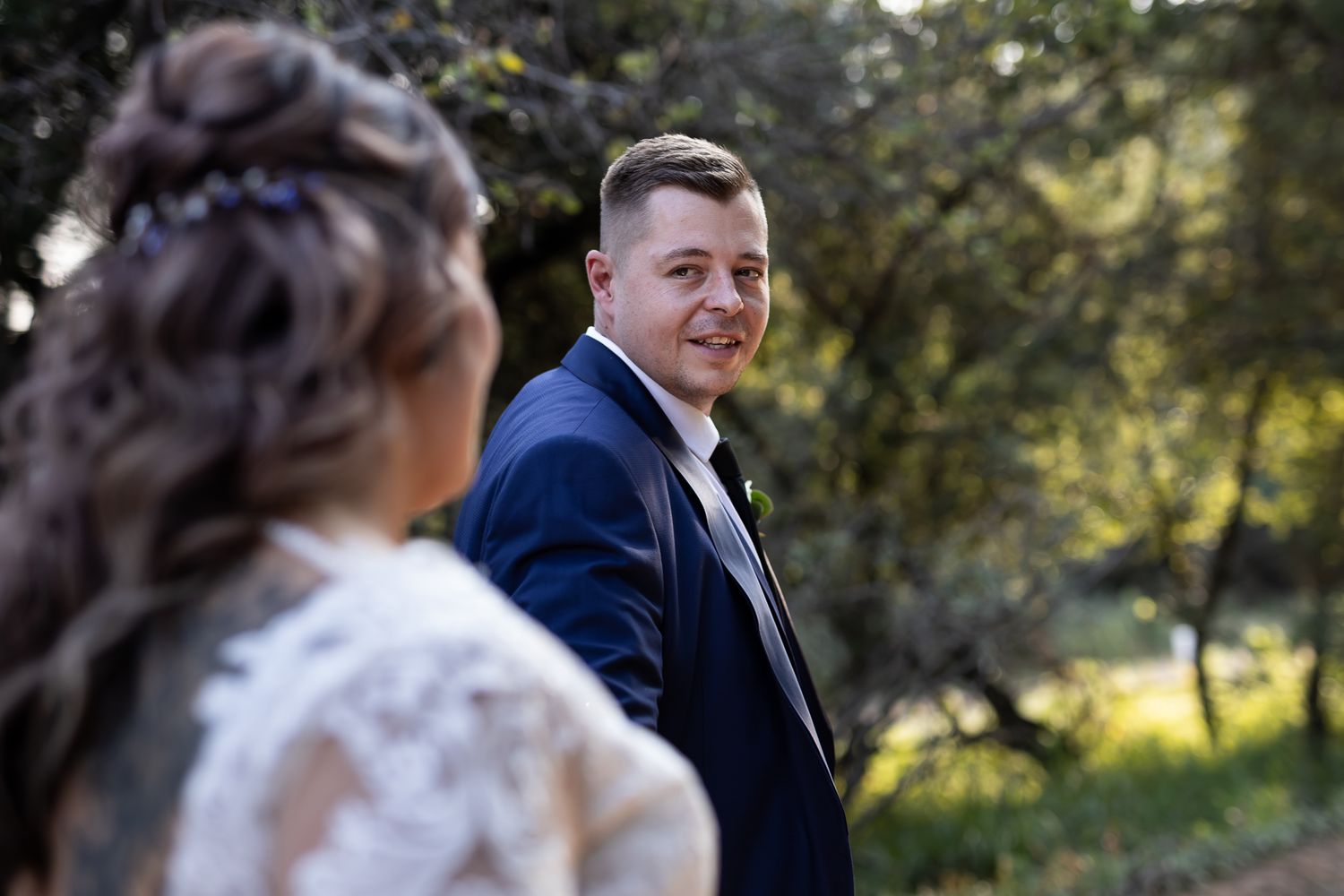 Groom in navy suit looks back at bride during outdoor wedding photoshoot.