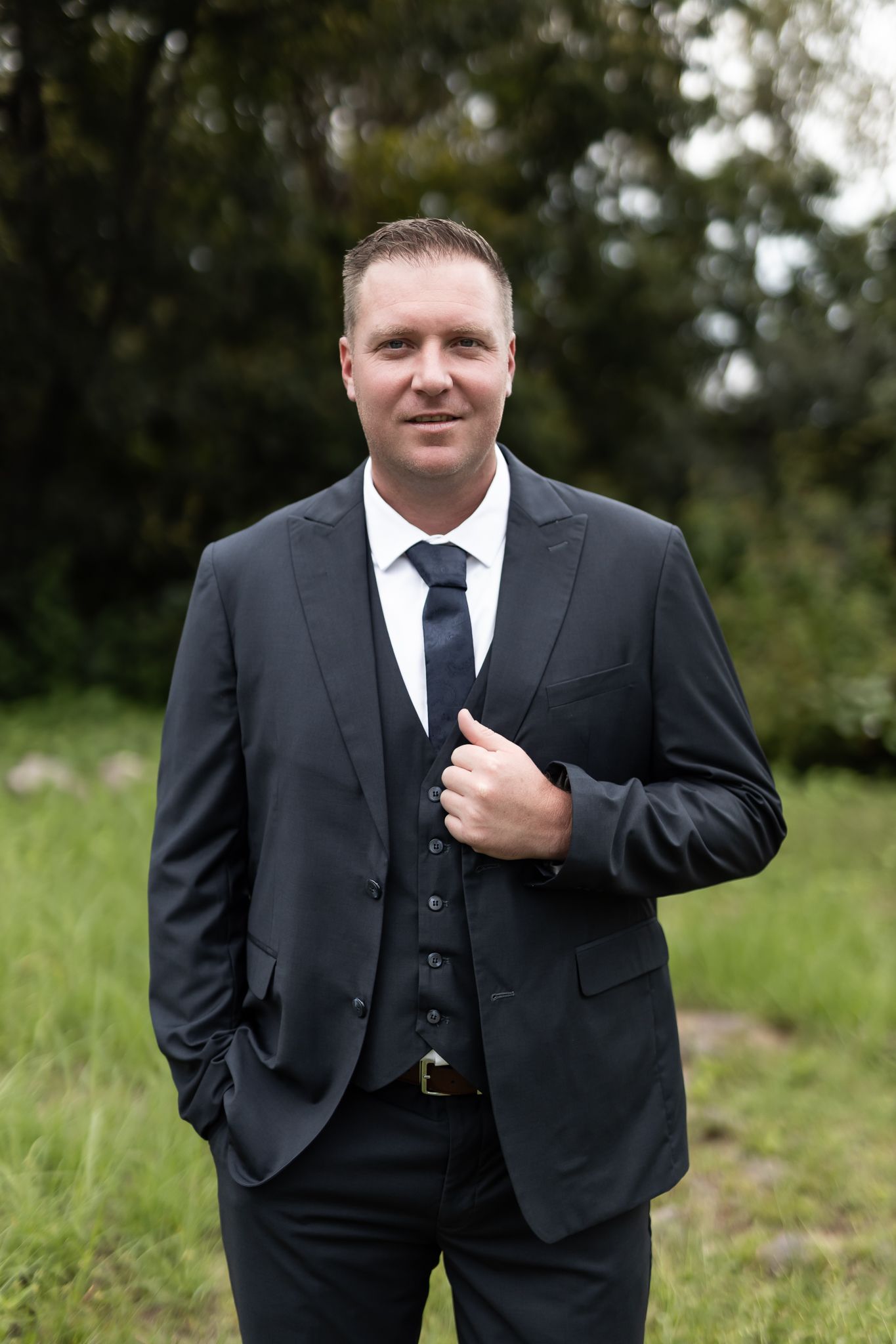 Groom in dark three-piece suit poses confidently against natural outdoor backdrop.