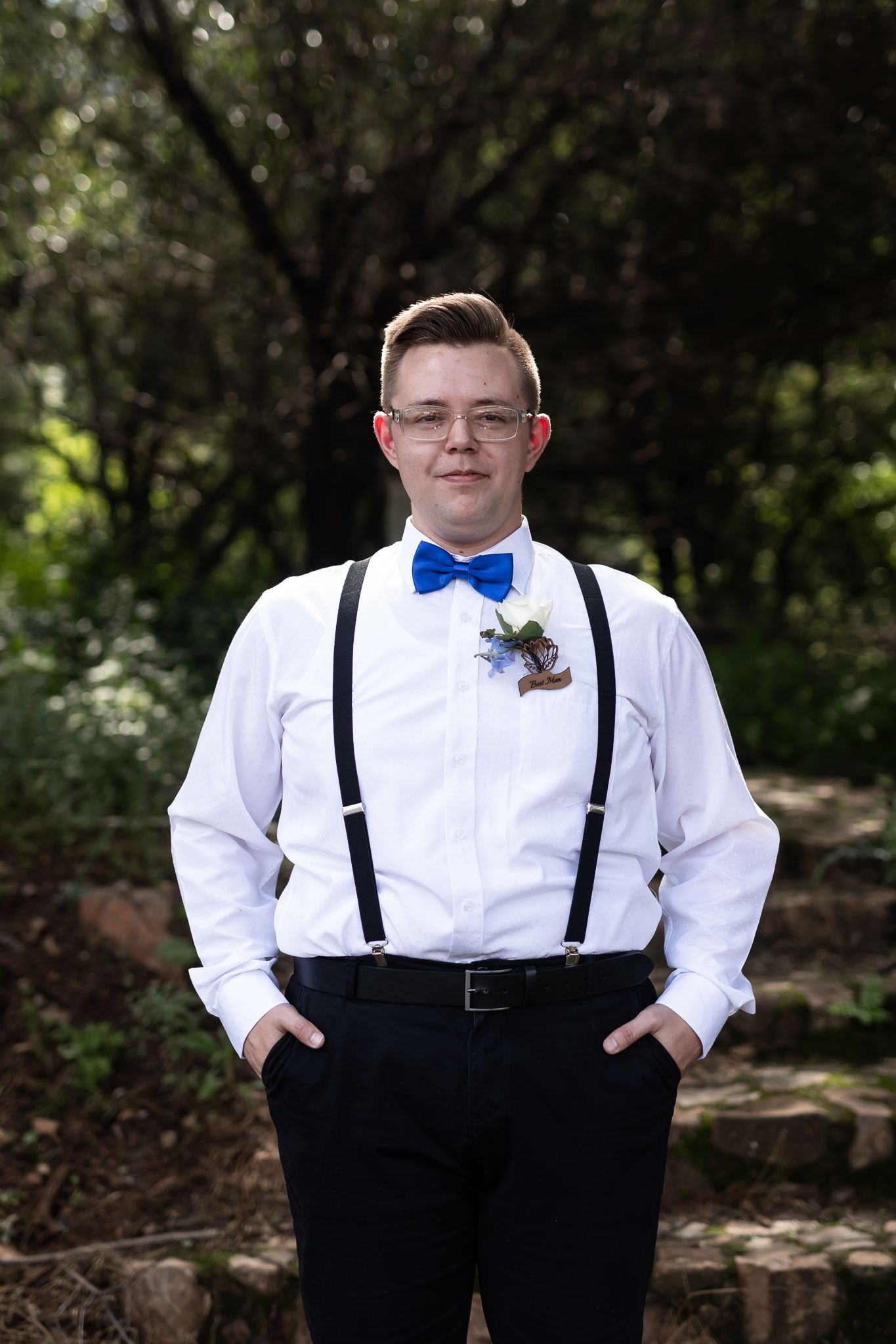 Groomsman in white shirt, suspenders and blue bowtie poses outdoors.
