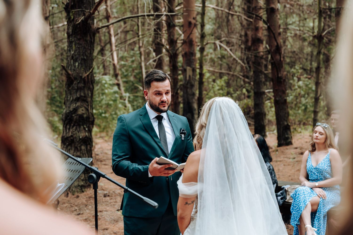 Couple exchanges vows during intimate outdoor forest wedding ceremony with guests watching in background.