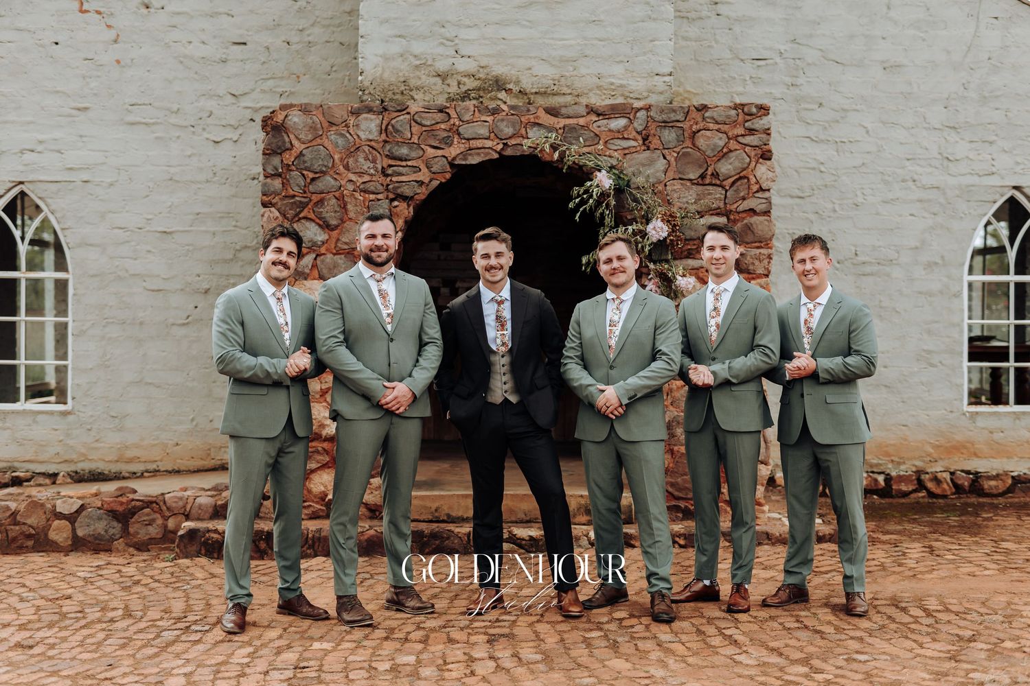 Wedding party in gray suits posed casually against rustic brick wall outdoors.