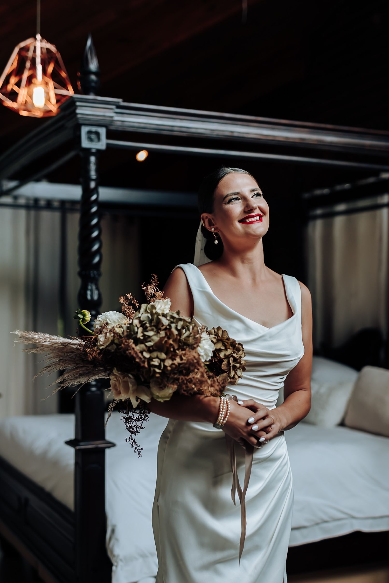 A bride in a sleek white wedding dress holds a rustic dried floral bouquet while standing in an elegant indoor setting.