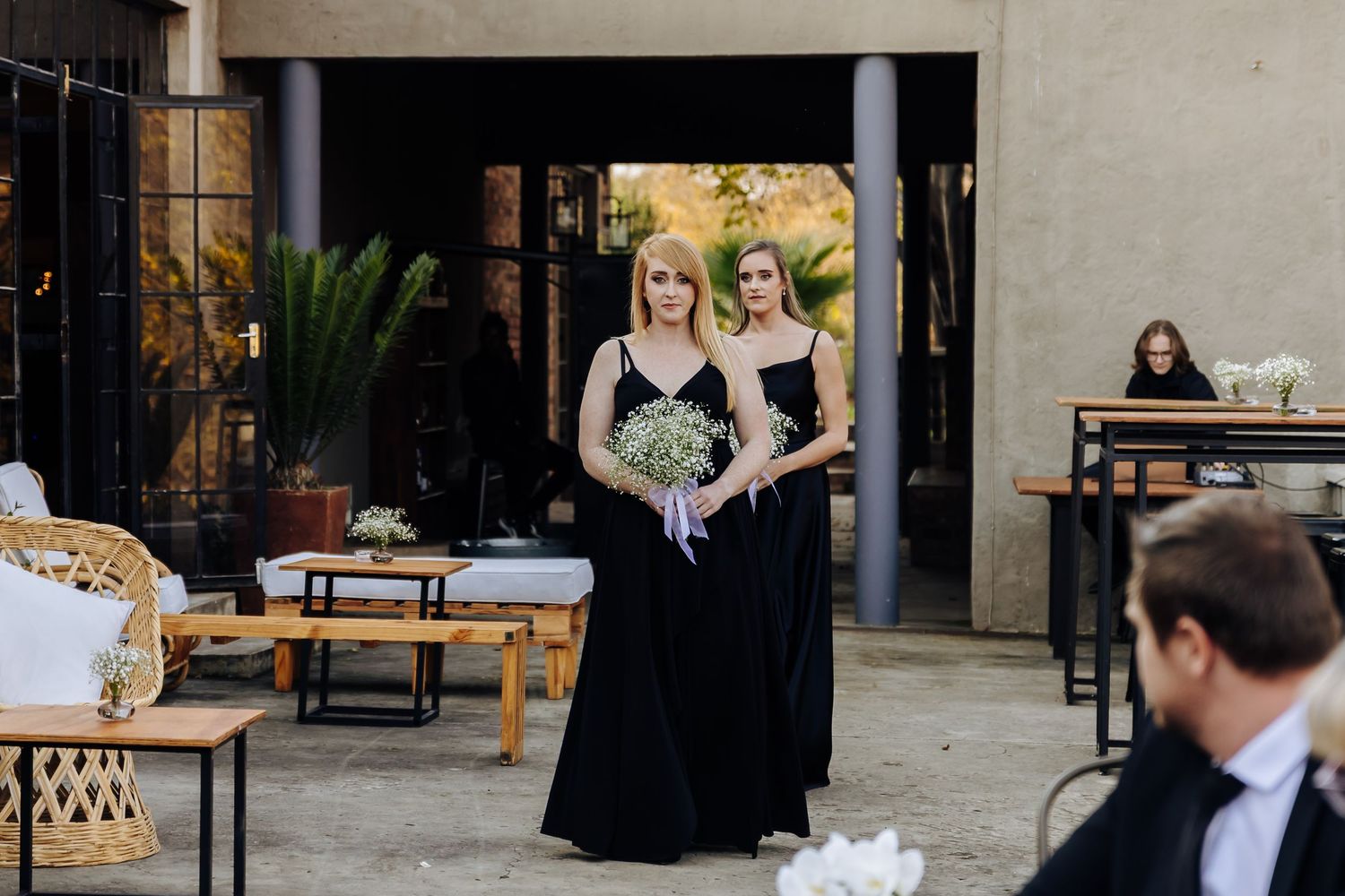 Two bridesmaids in elegant black gowns stand at an outdoor wedding ceremony venue with patio furniture visible.