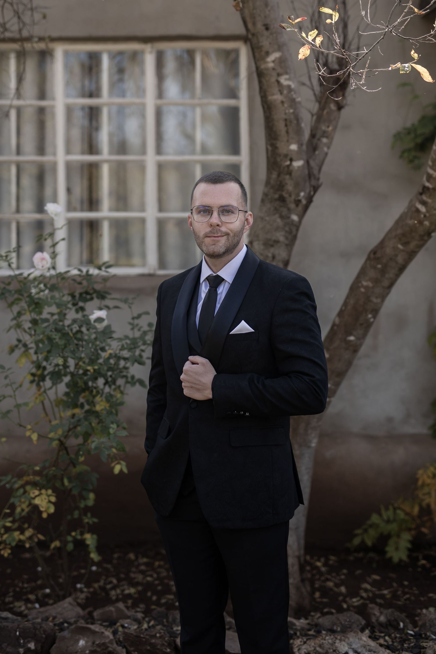 Professional portrait of someone in a black suit and glasses standing outdoors next to a building with windows.